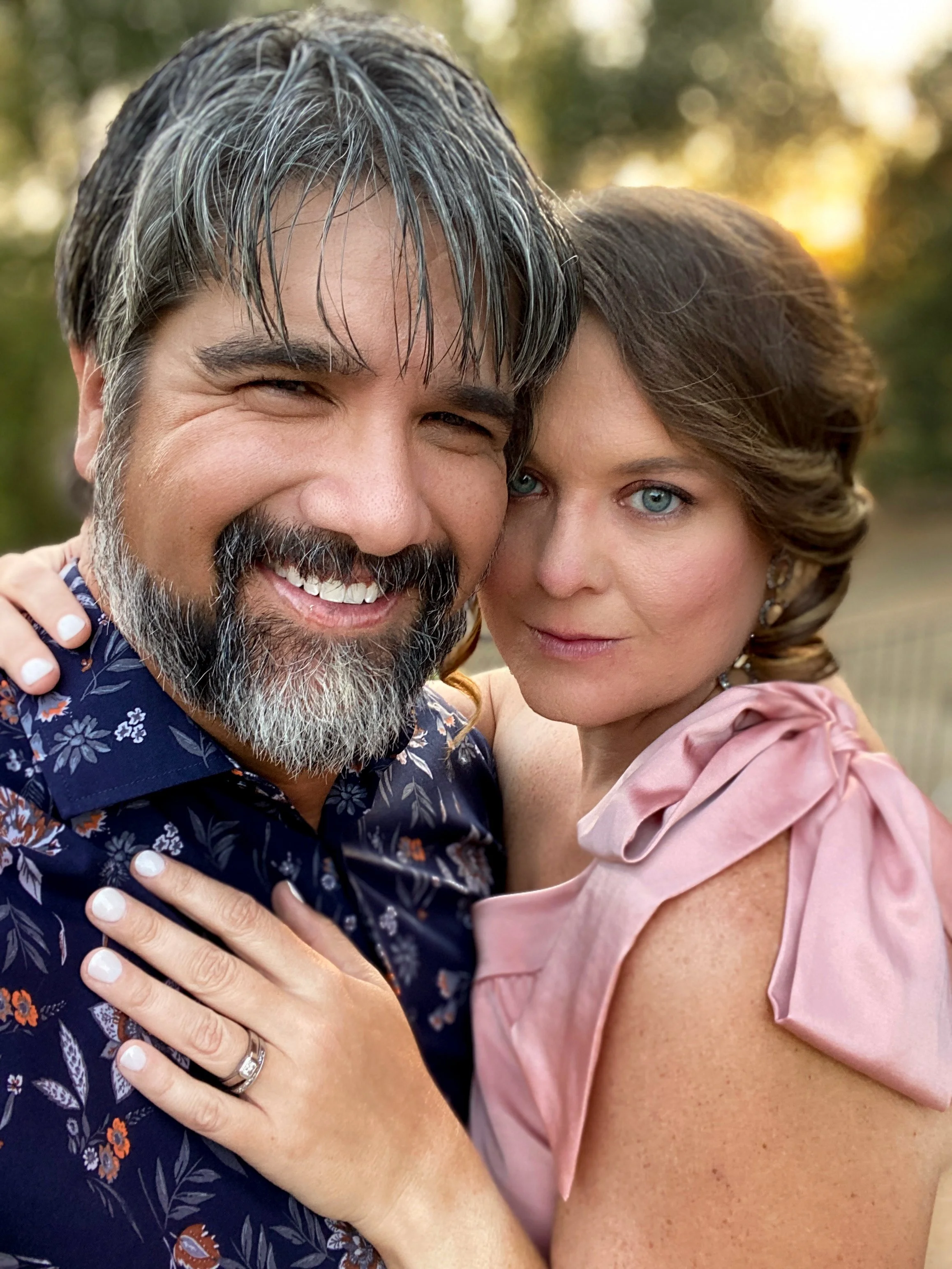 A man with gray hair and beard smiling, and a woman with blue eyes and brown hair, close together outdoors during sunset, wearing floral and pink dresses.
