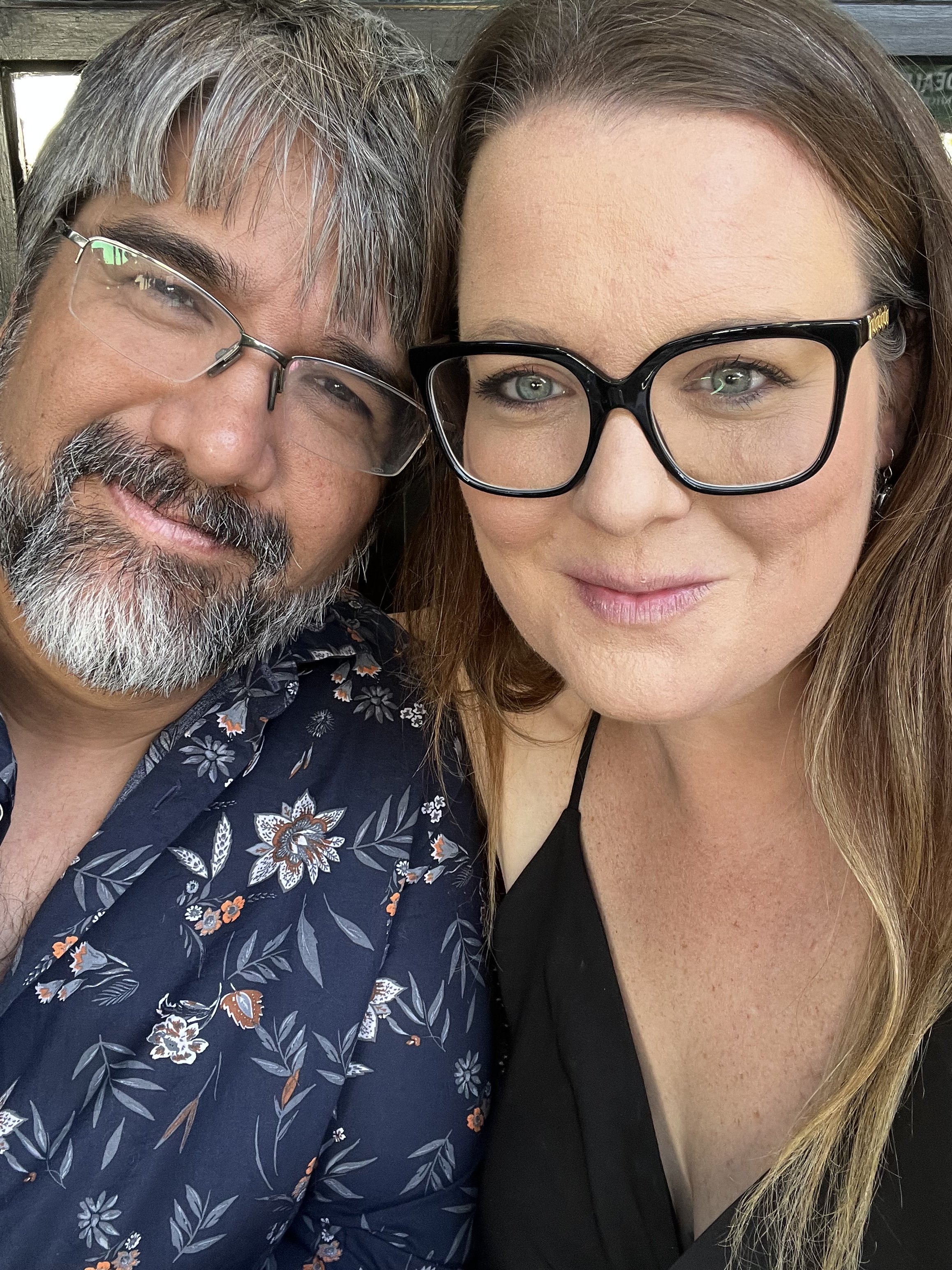 A middle-aged man with gray hair, a beard, and glasses smiling next to a woman with long, light brown hair, green eyes, and black glasses, both close-up.