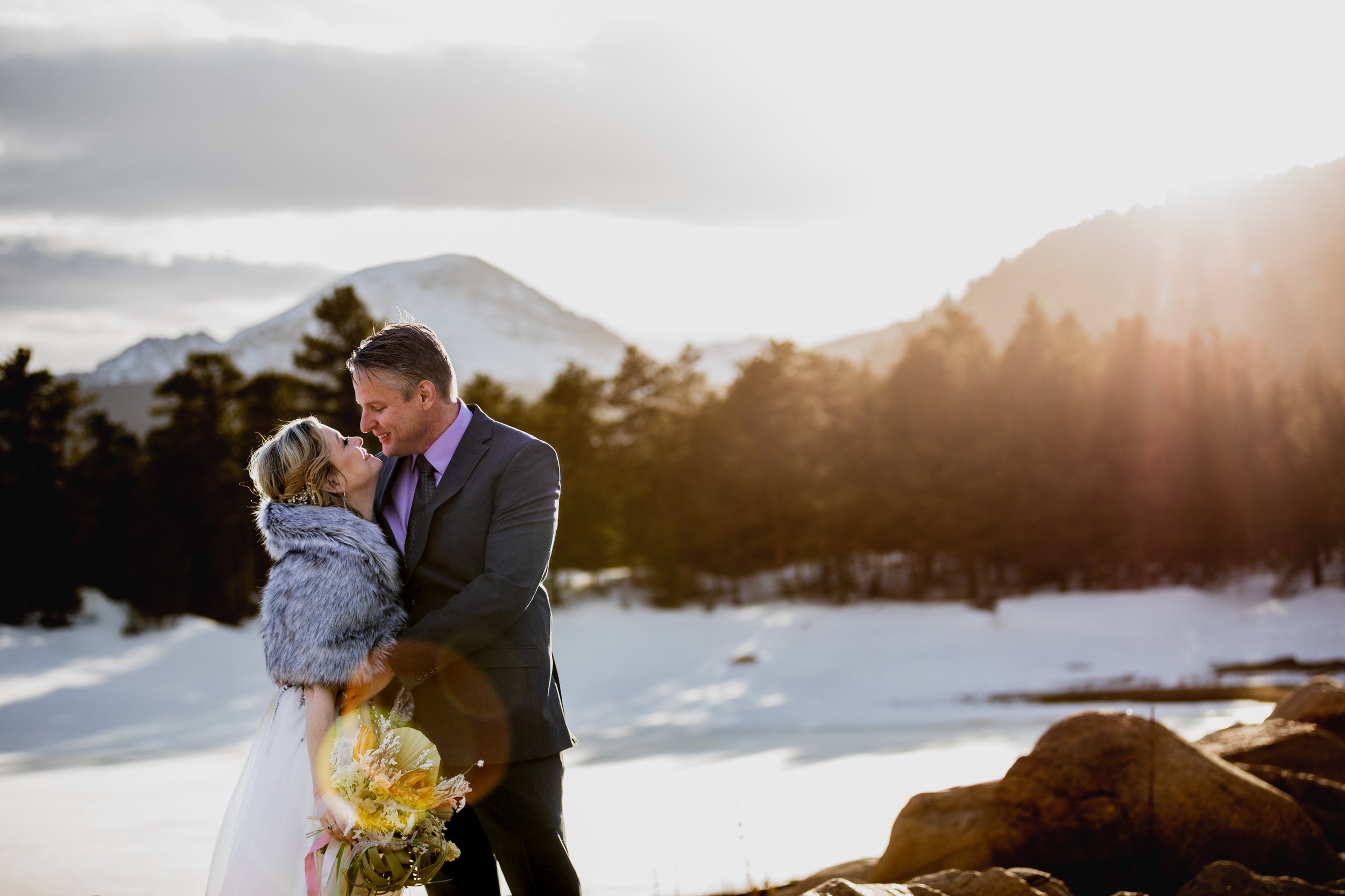 A couple in wedding attire sharing a romantic moment outdoors in a snowy mountainous landscape at sunset.