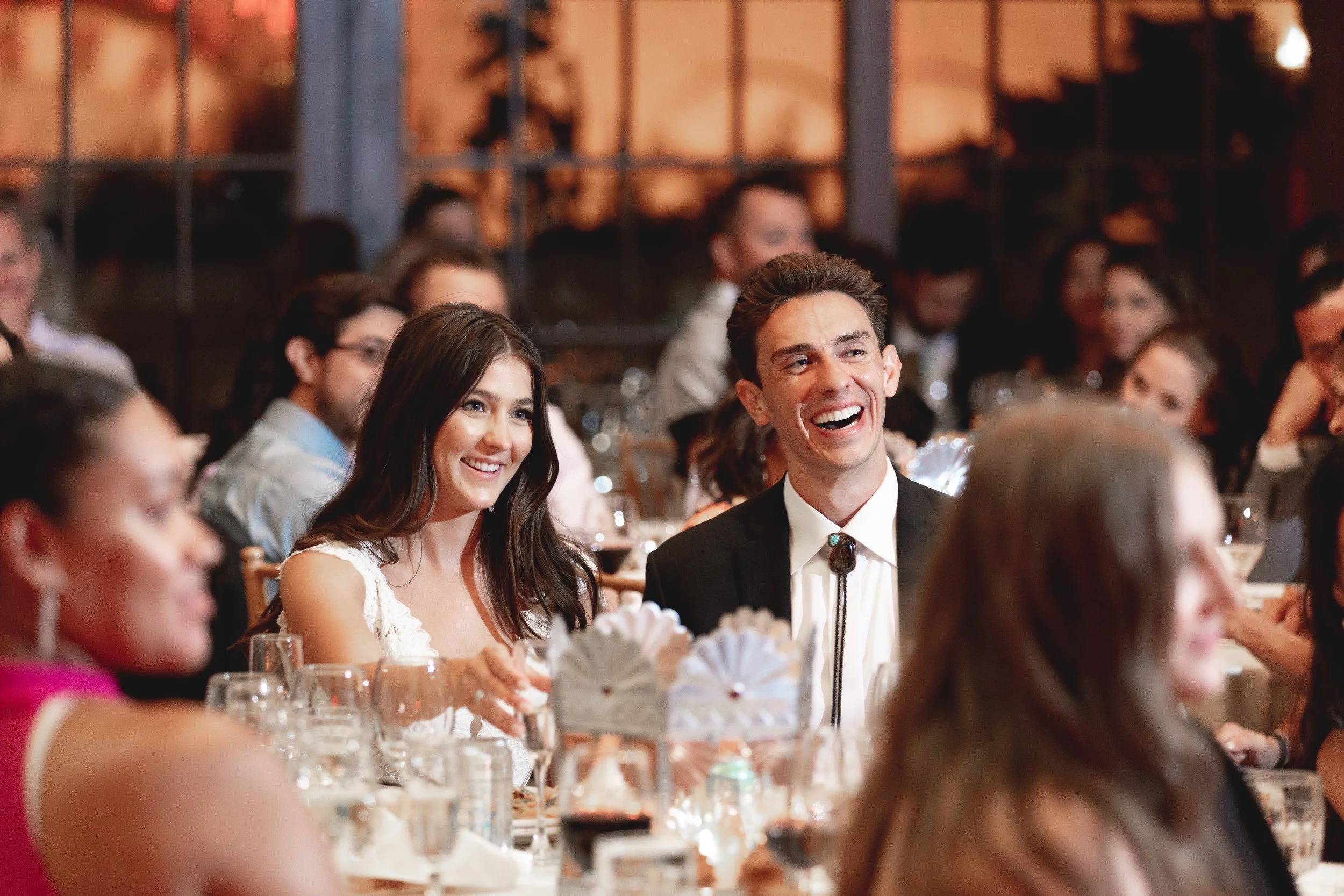 People attending a formal event, dressed in elegant attire, seated at a table with glasses and napkins, smiling and enjoying socializing.