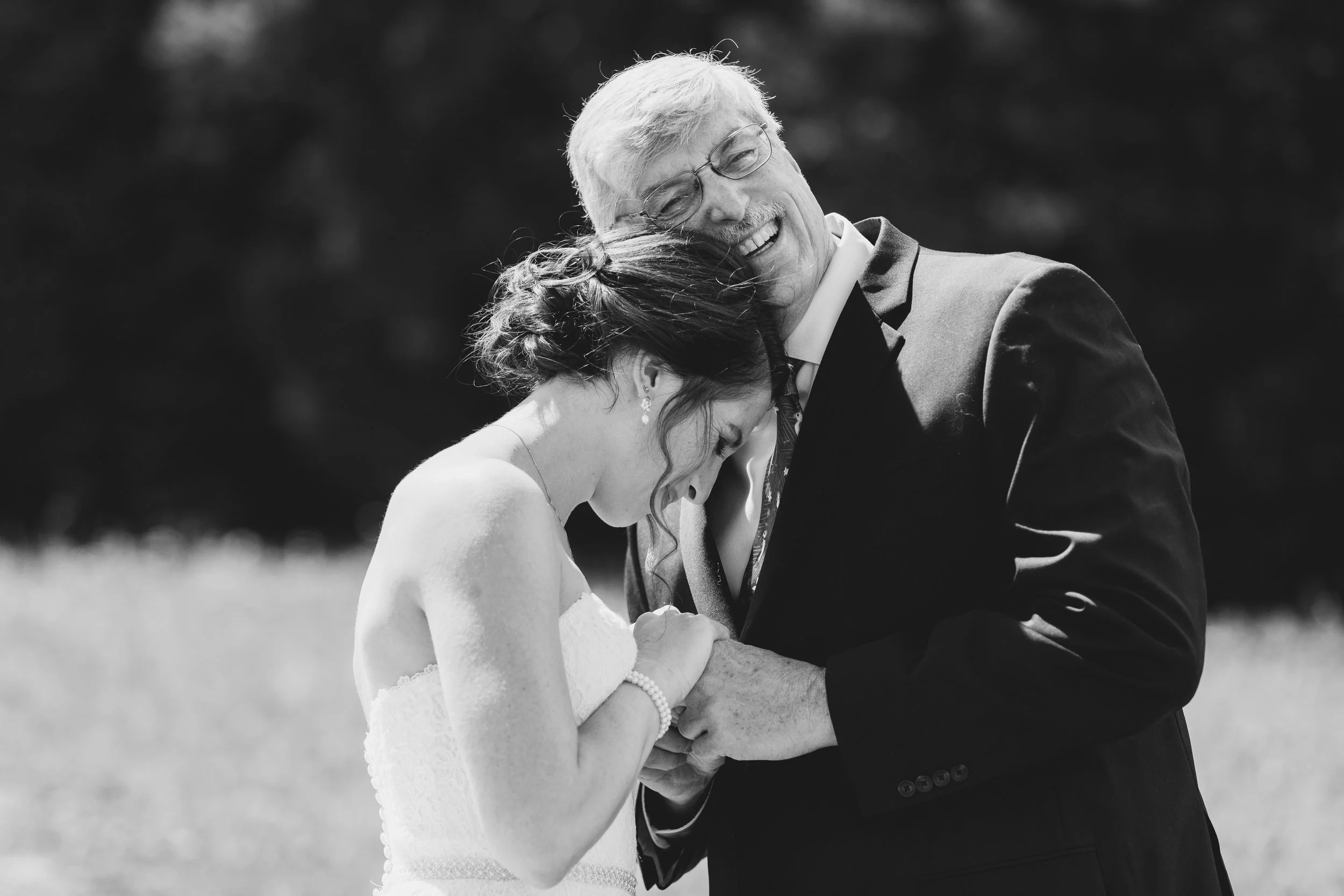 An elderly man in a suit embracing a young woman in a wedding dress, both with their heads bowed together, sharing an emotional moment outdoors.