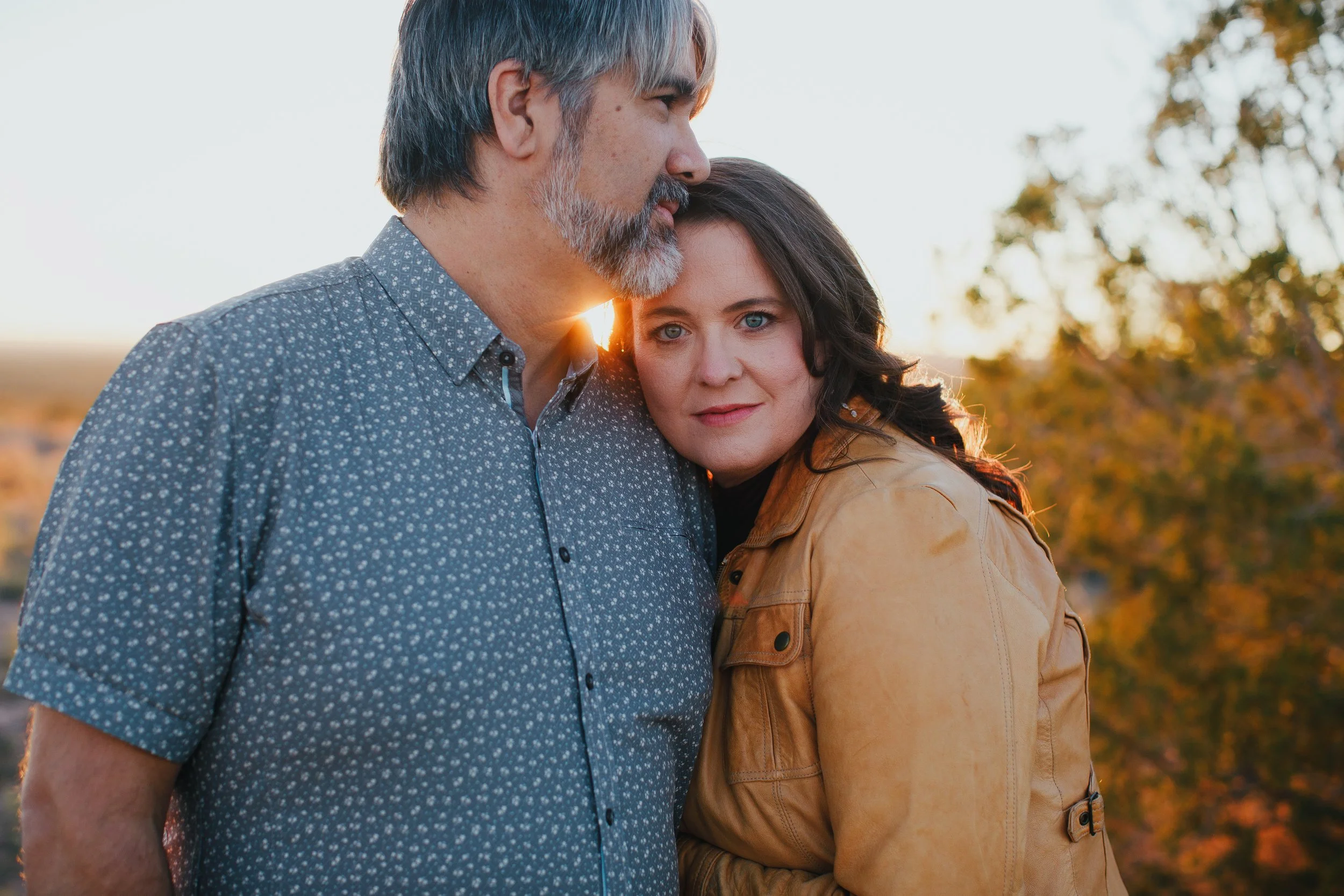 A middle-aged man with gray hair and beard and a young woman with dark hair in an outdoor setting during sunset, standing close together, with the man resting his forehead on the woman's head.