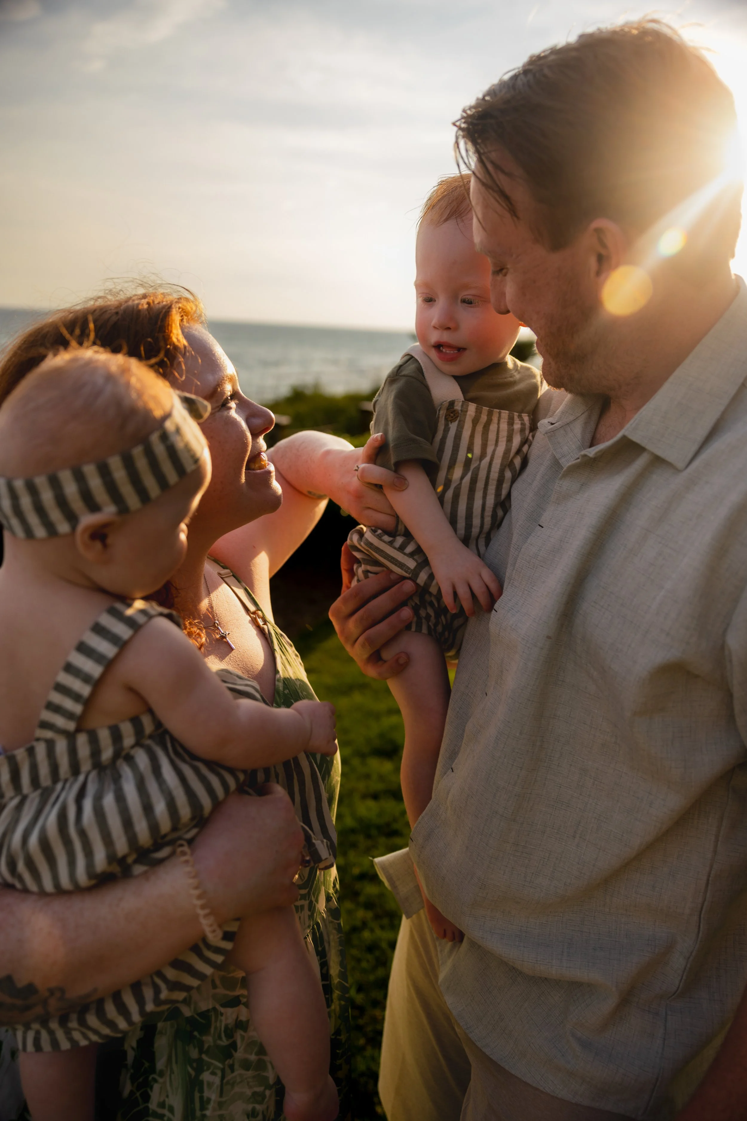 A family of four at sunset by the beach, smiling and playing together. The mother and father holding and engaging with two young children, all smiling.