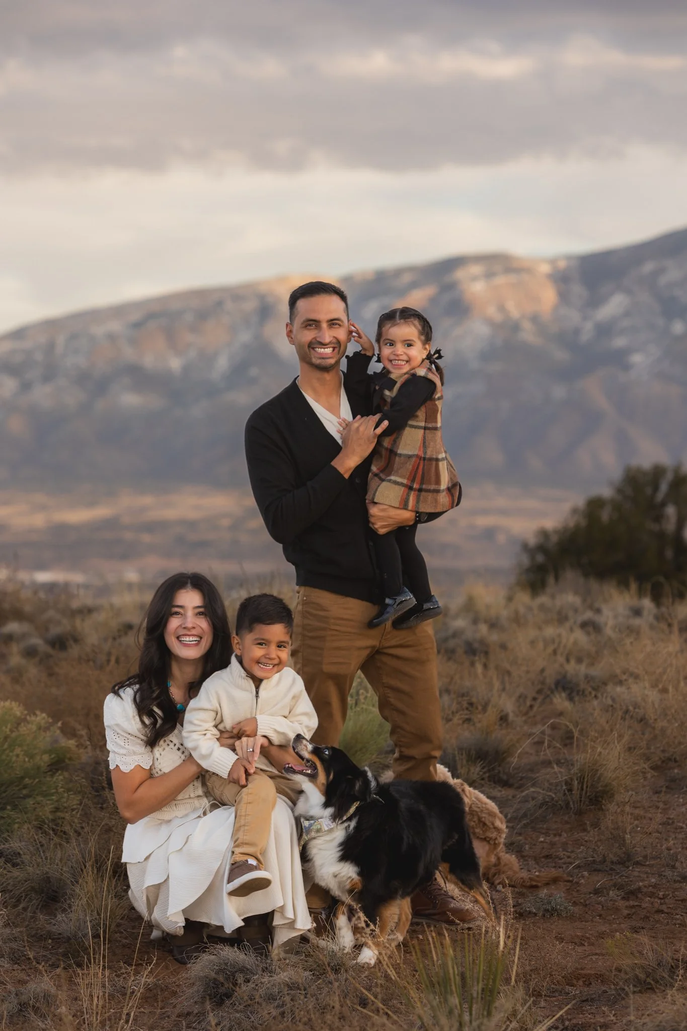 A happy family of four with a dog outdoors in a desert landscape with mountains and cloudy sky.