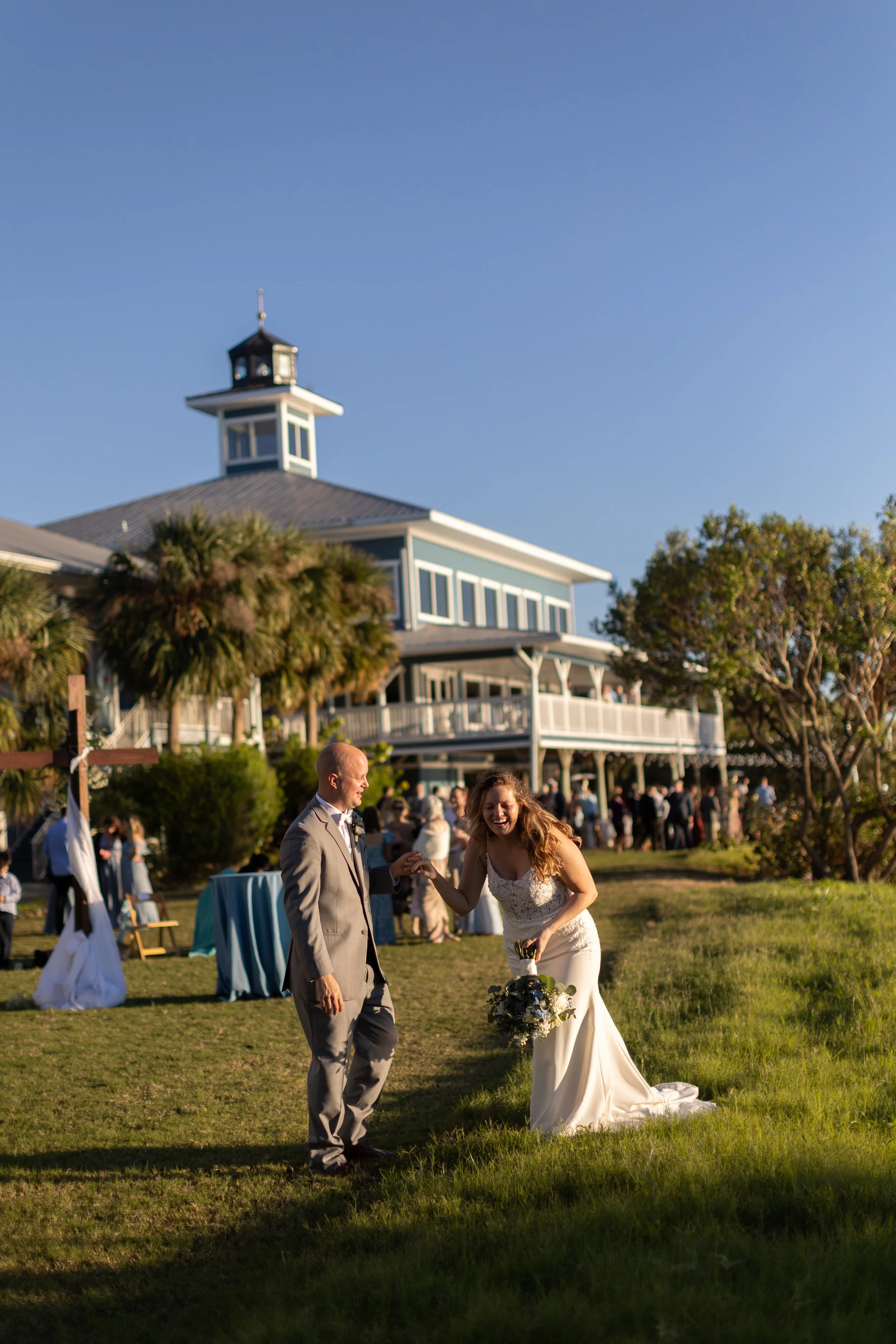 A bride in a white wedding dress laughing and dancing with an older man in a suit outdoors on a sunny day, near a large house or venue with a tower, trees, and a gathering of people in the background.