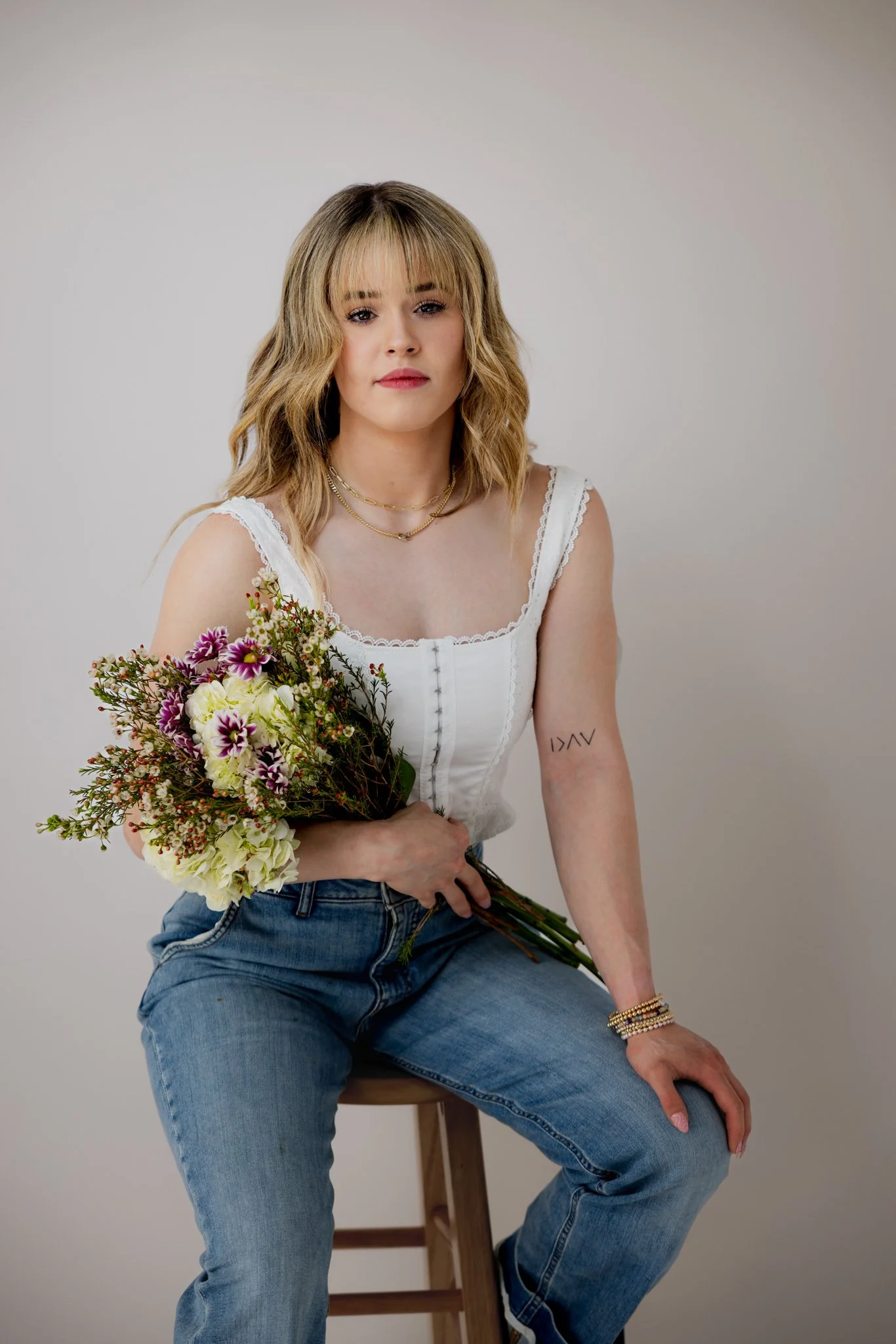 Young woman with shoulder-length hair holding a bouquet of flowers, wearing a white top and jeans, sitting on a wooden stool against a plain background.
