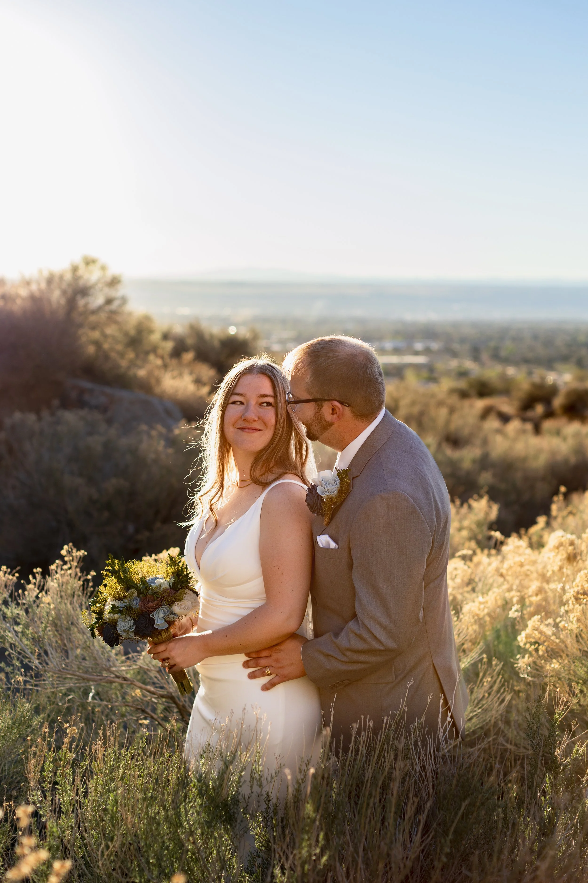 A bride and groom standing close together outdoors in a scenic, natural landscape during sunset. The bride is holding a bouquet of flowers and smiling softly, while the groom is gently kissing her on the cheek. The background features rolling hills, shrubs, and a clear sky.