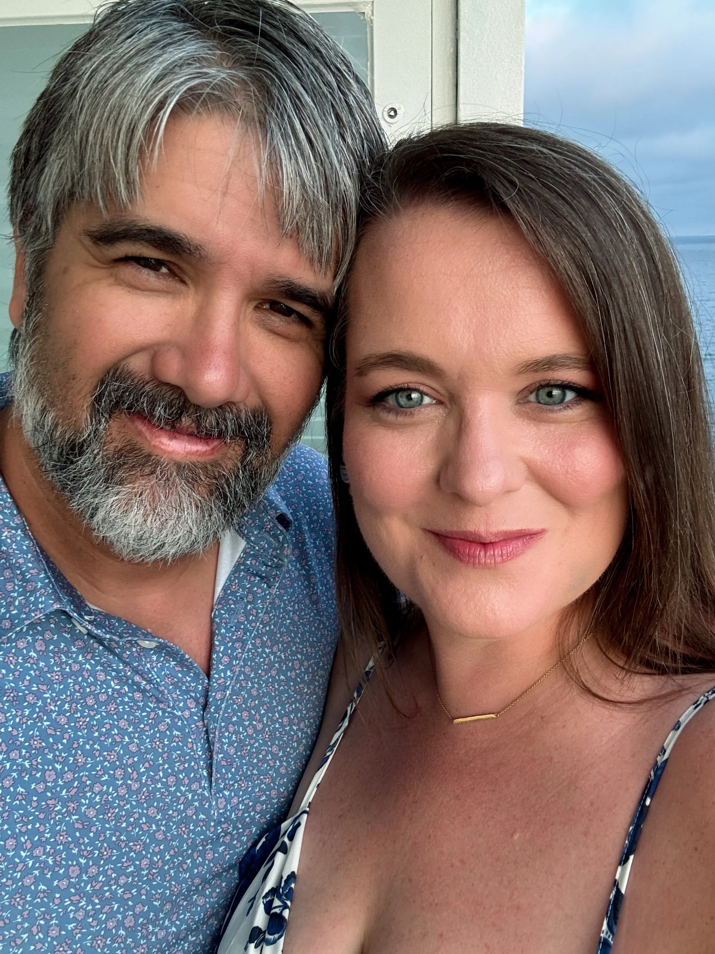 A close-up of a smiling middle-aged man with gray hair and beard, and a smiling woman with blue eyes and brown hair, standing together outdoors with a blurred ocean background.