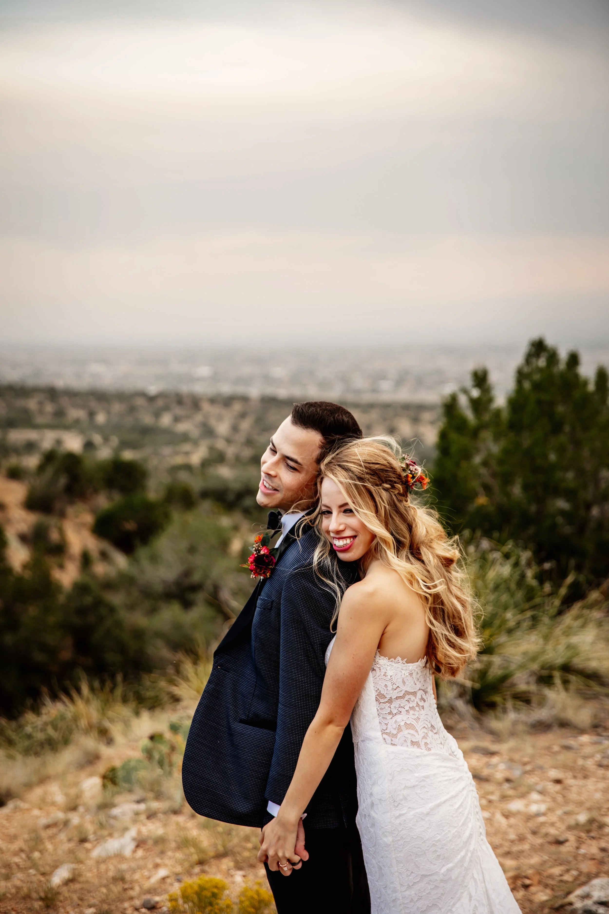A smiling bride and groom stand close together outdoors with a scenic landscape in the background, during their wedding photoshoot.