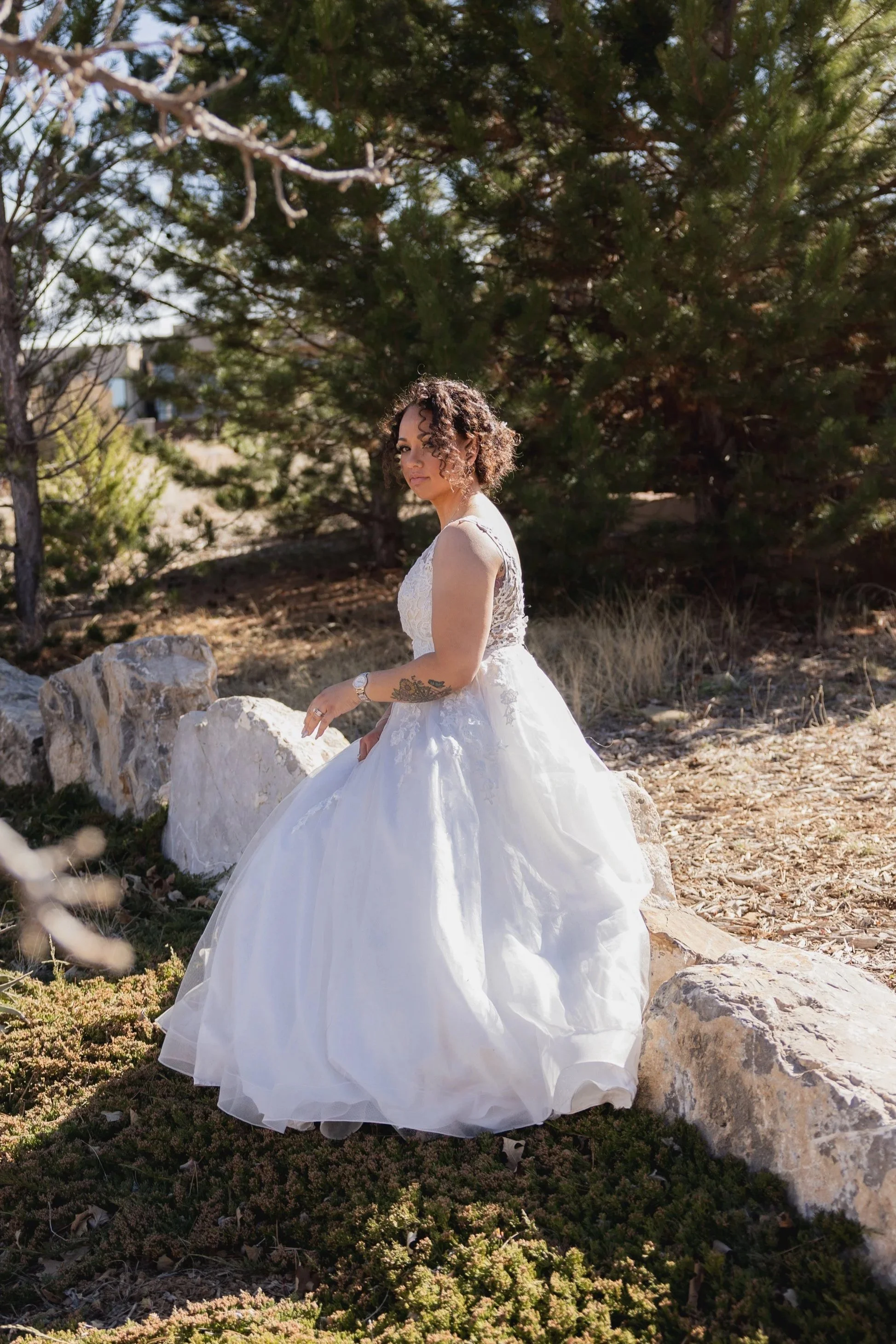 A woman in a white wedding dress sitting on rocks outdoors among trees.