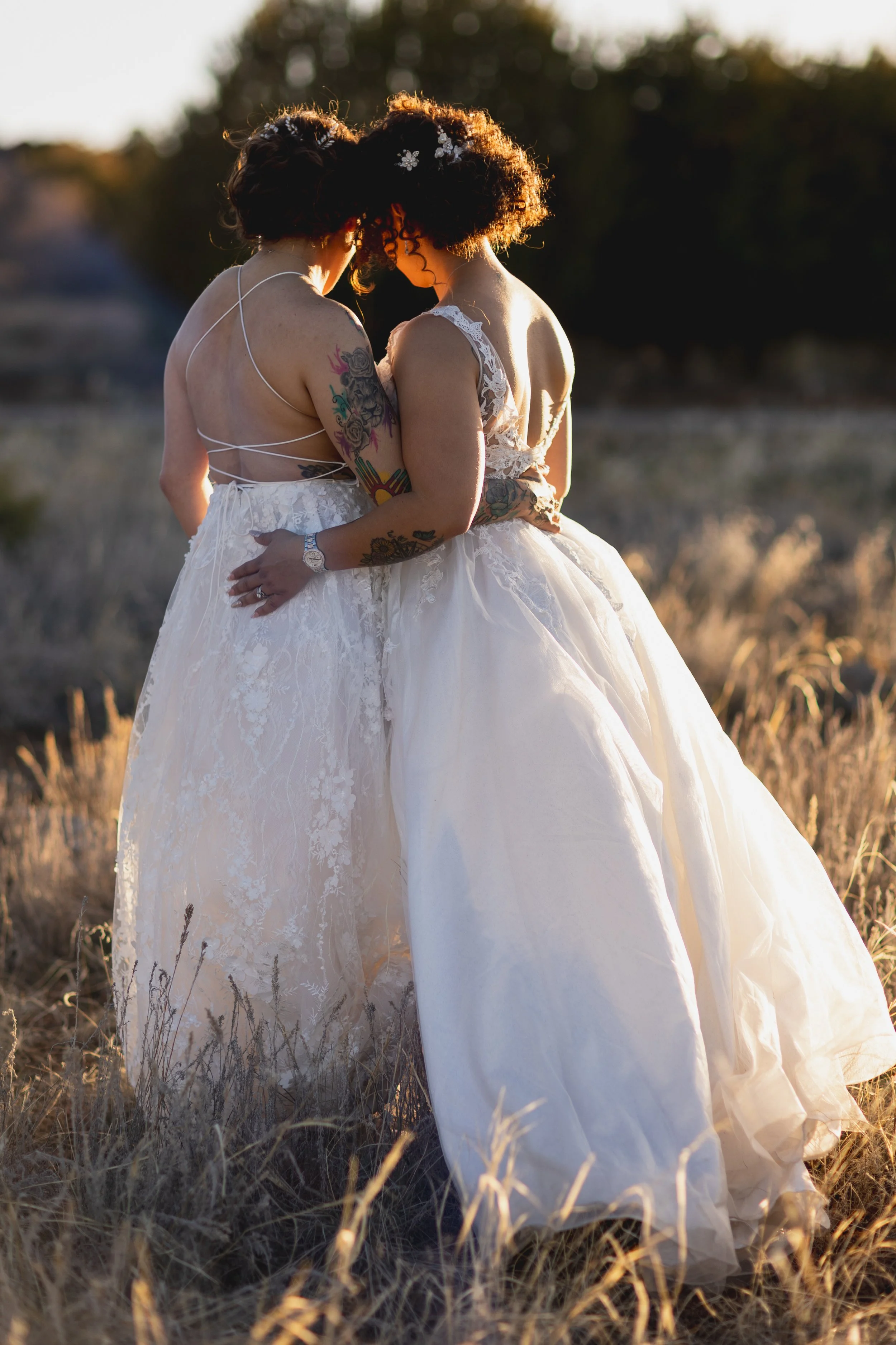 Two women wearing wedding dresses standing close together in a field at sunset, their foreheads touching and embracing.