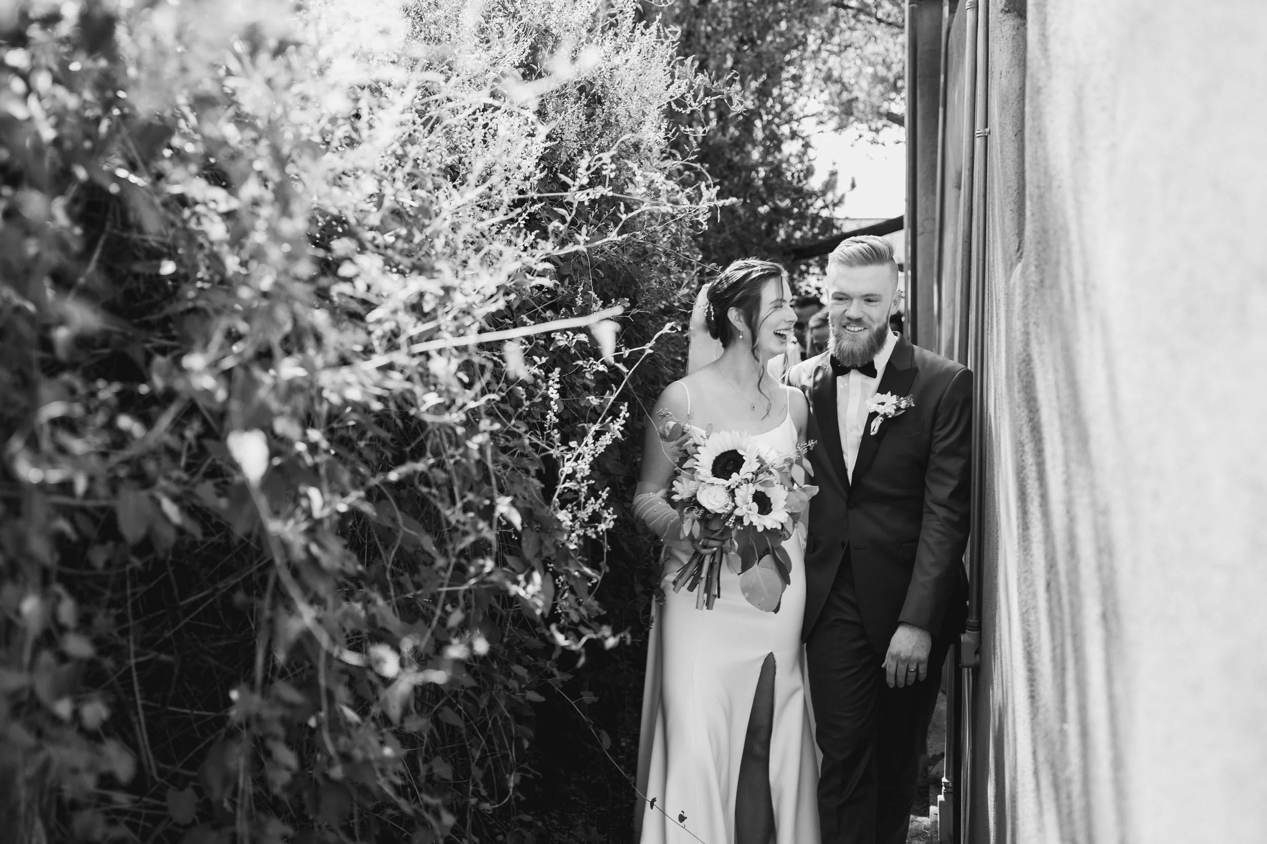 Black and white photo of a bride and groom smiling and walking together outdoors. The bride is holding a bouquet and wearing a wedding dress with a slit, while the groom is in a tuxedo. They are close to a wall and surrounded by foliage.