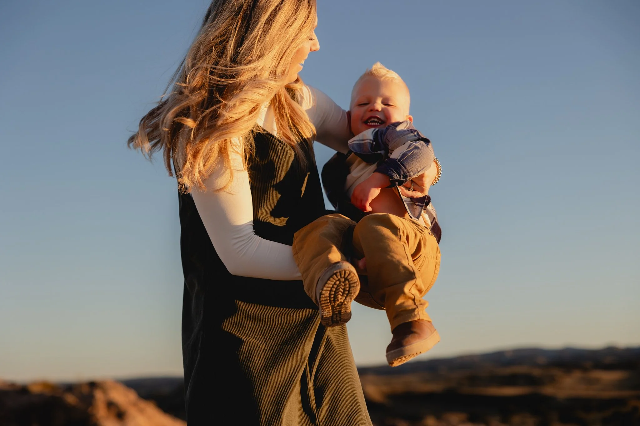 A woman with long, wavy blonde hair holding a smiling young boy with blond hair in her arms outdoors during sunset with a clear sky and landscape in the background.