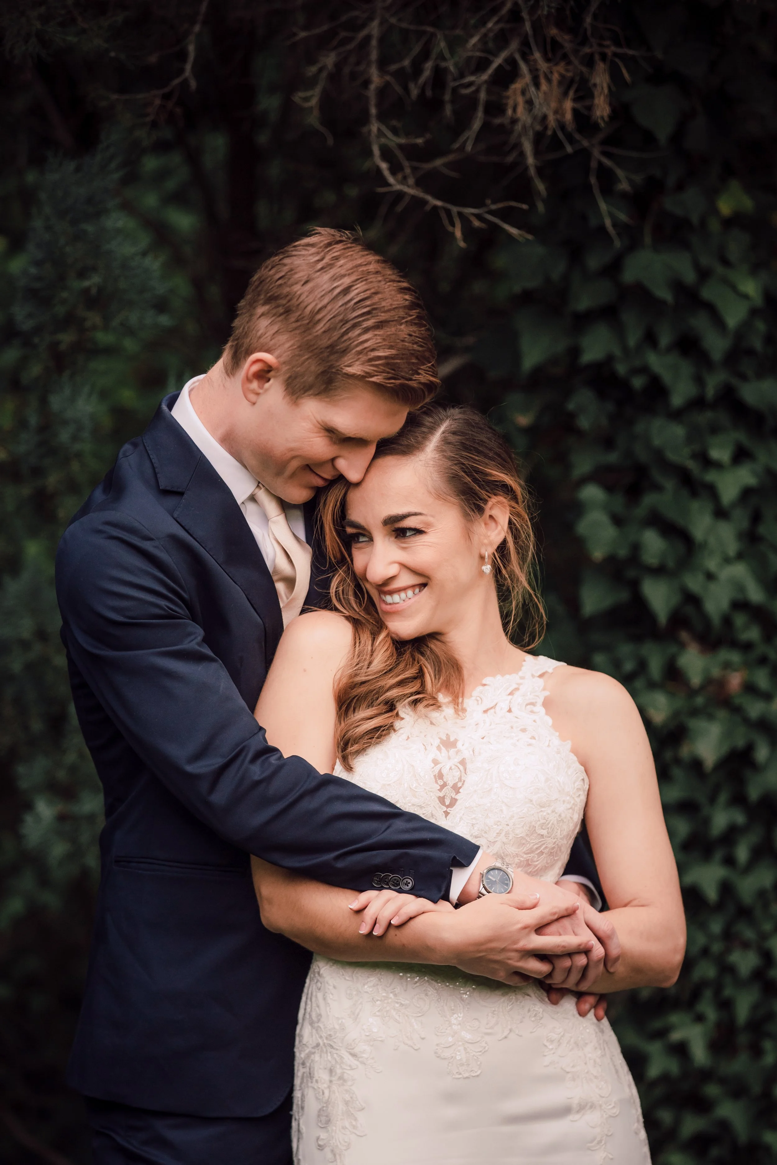 A groom and bride embracing outdoors, the groom leaning his forehead against the bride's head, both smiling, with greenery in the background.