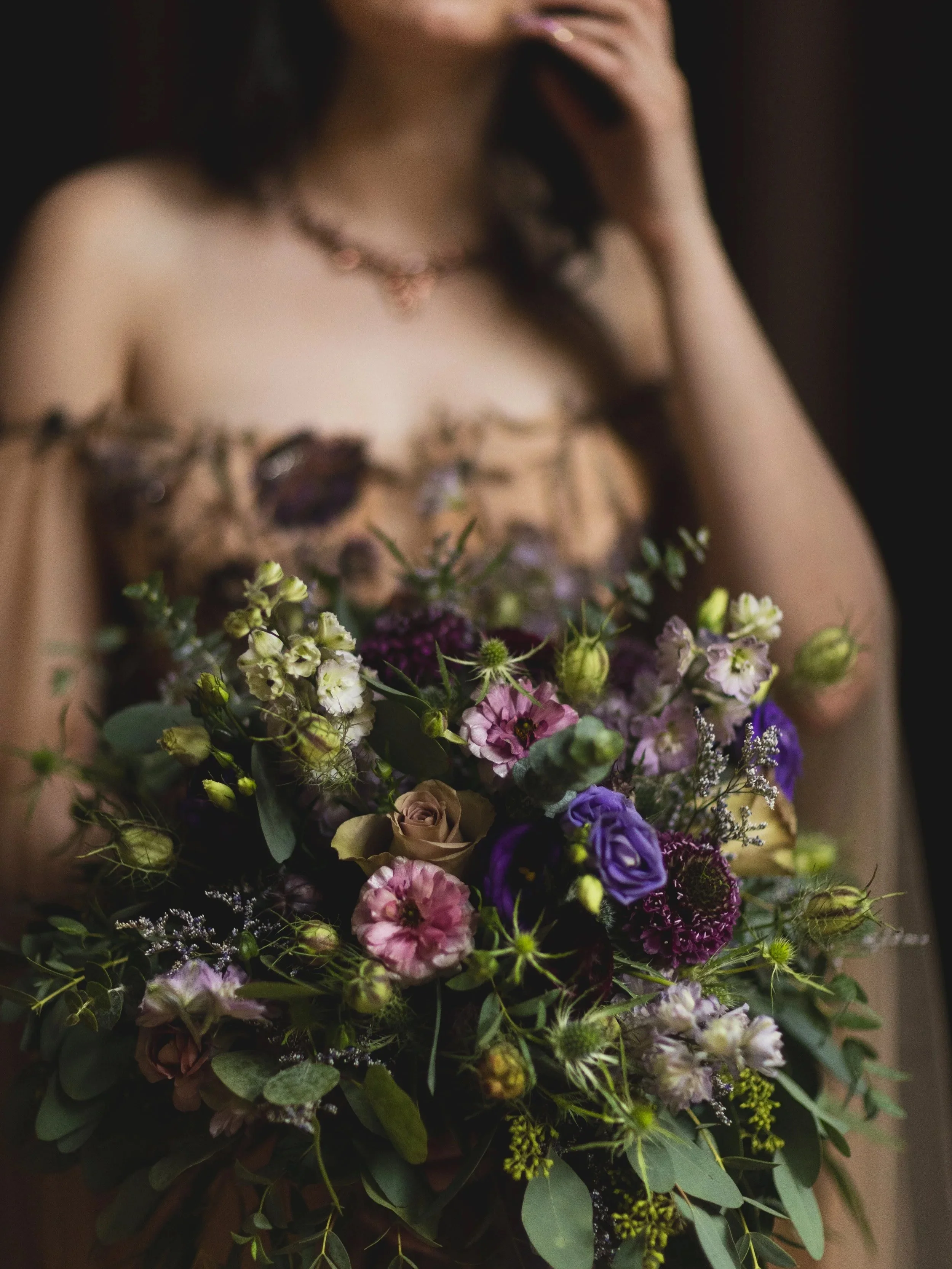 Woman holding a bouquet of mixed purple, pink, white, and cream flowers, wearing a floral dress and necklace, with her face partially visible.