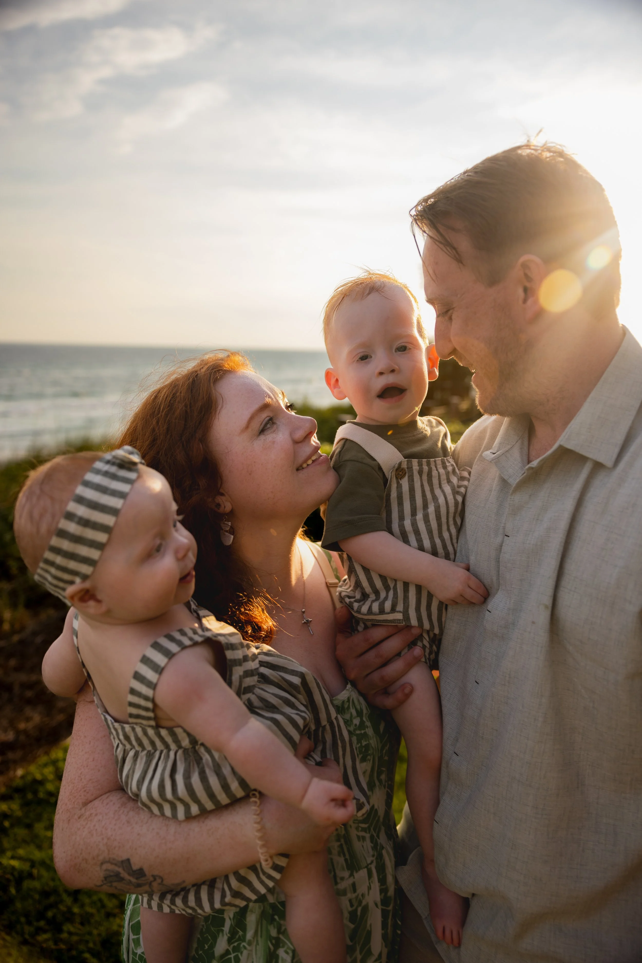 A family of four at the beach during sunset, with the parents smiling at each other and their two young children, all wearing matching striped outfits.
