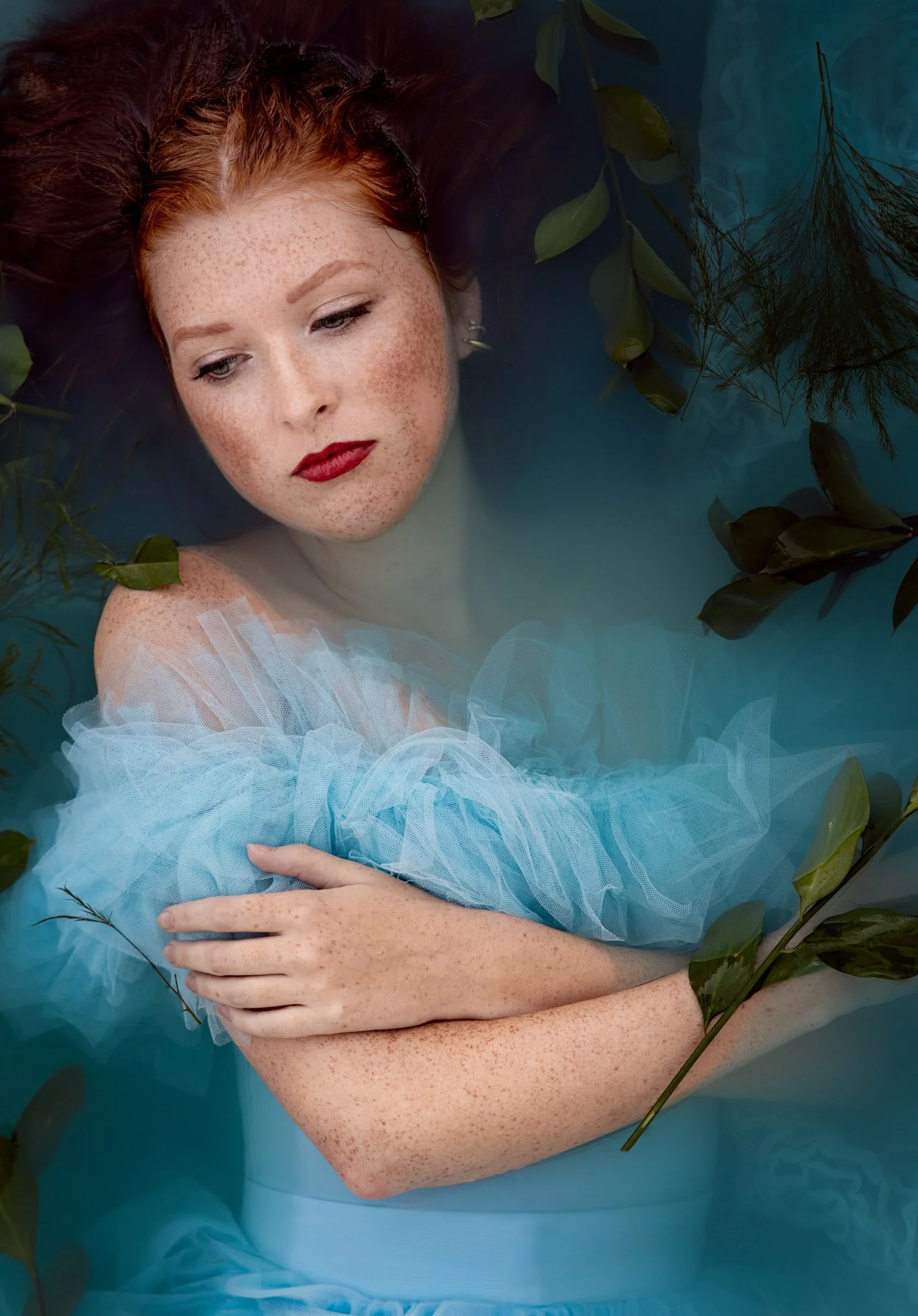 A red-haired woman with freckles submerges in a blue-tinted water scene, wearing a light blue tulle dress, surrounded by green leaves, with a serene expression.