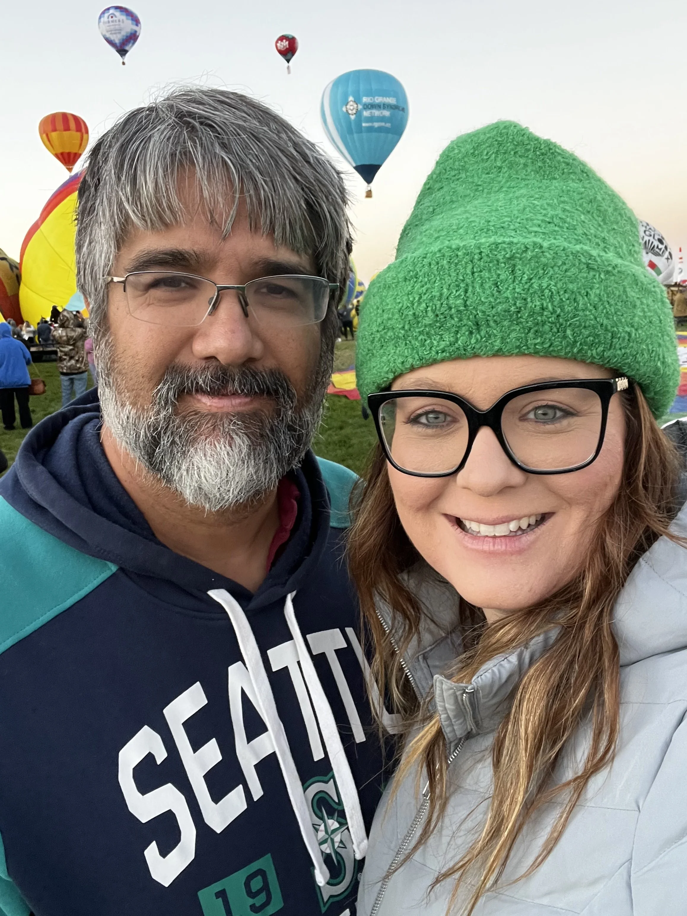 A man with gray hair, a beard, and glasses, and a woman with glasses and a green knit hat, smiling at a hot air balloon festival with colorful hot air balloons in the background.