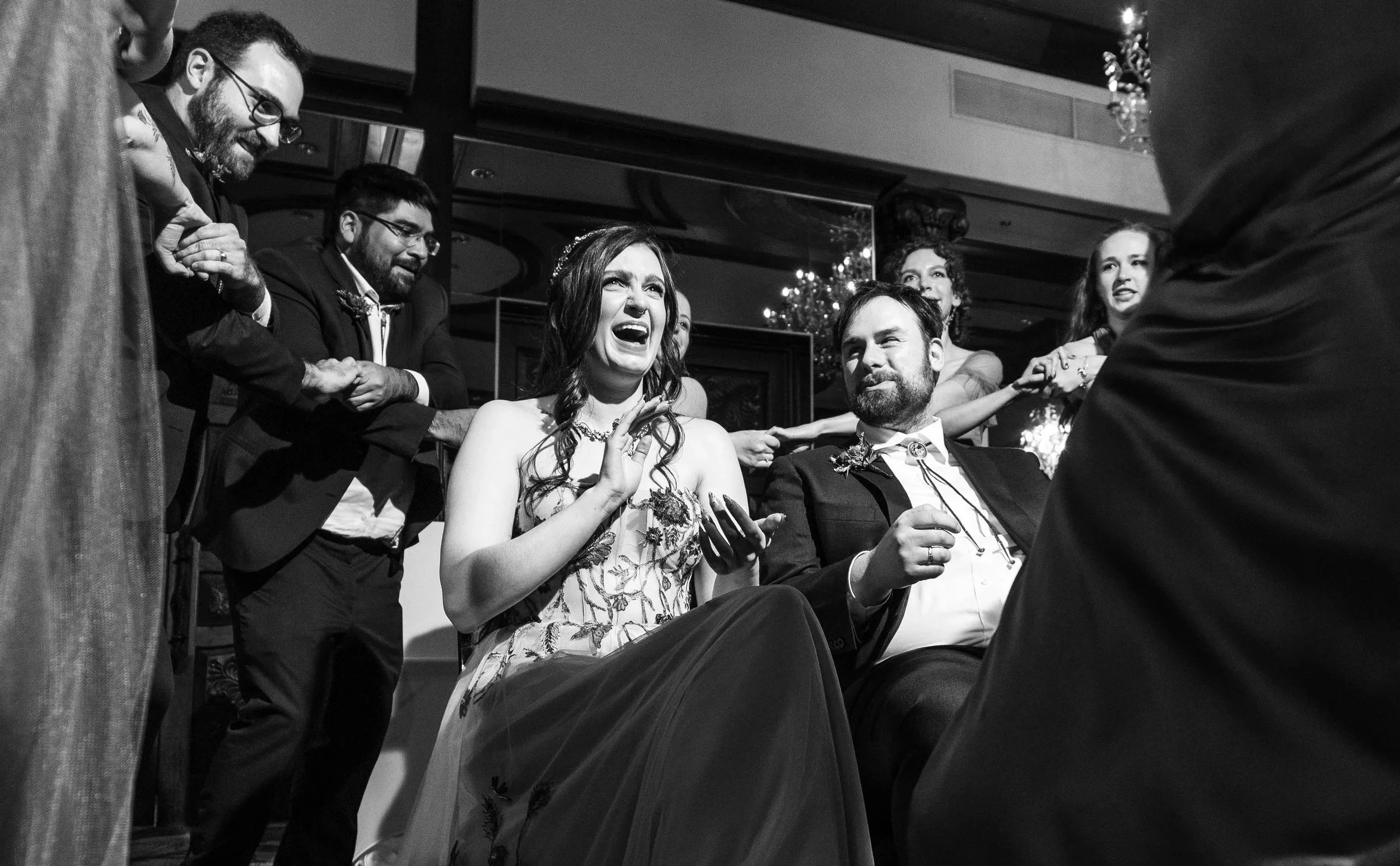 Black and white photo of a wedding scene with a woman smiling and a man sitting beside her, surrounded by guests holding hands, in an indoor venue.