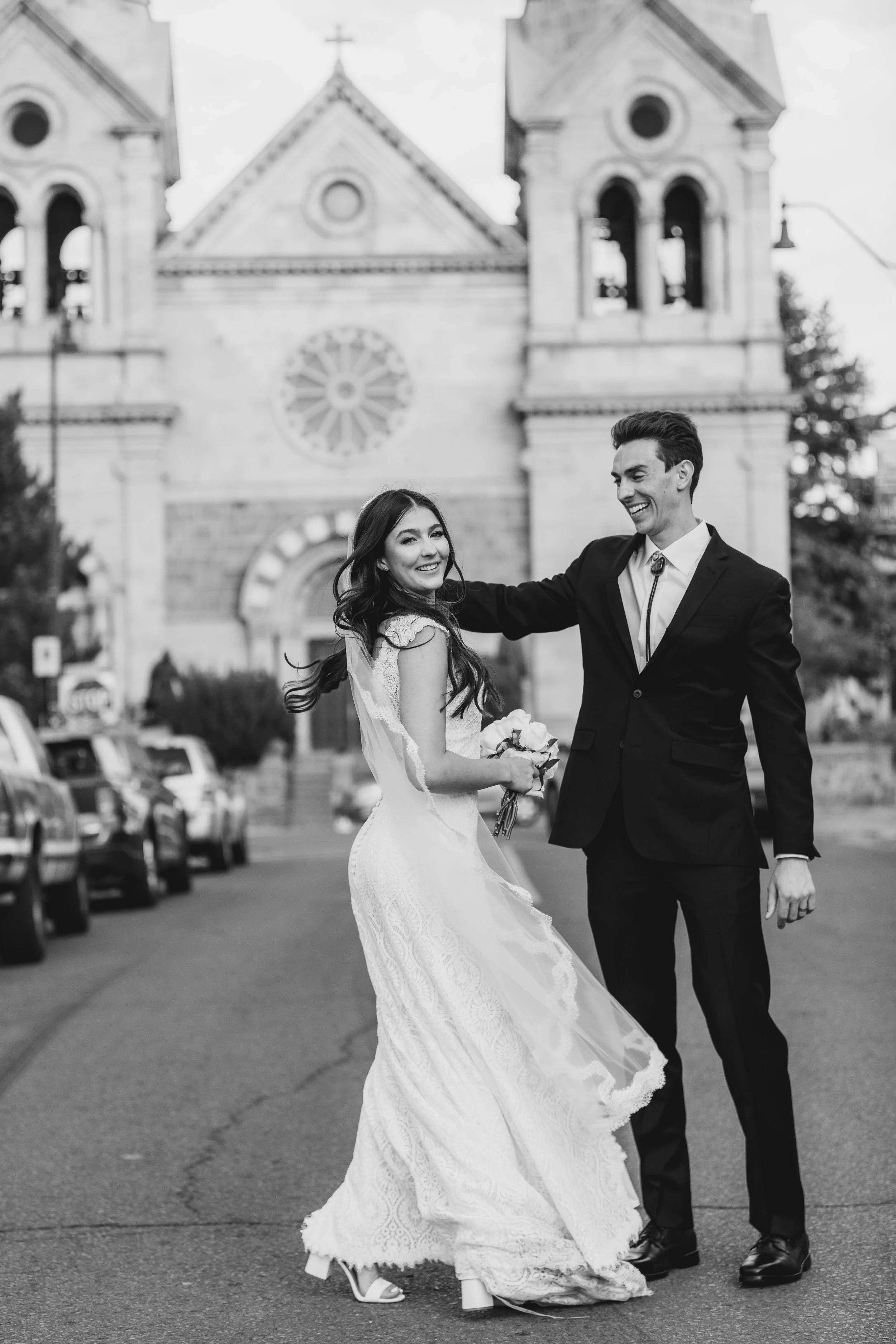 A black and white photo of a bride and groom smiling and dancing outside a church, with cars parked along the street in the background.