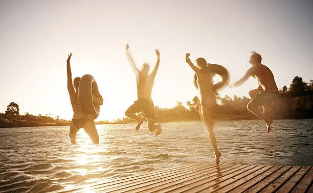 A joyful group of friends with inner tubes simultaneously jump into the sparkling, sun-drenched waters of Table Rock Lake, Missouri, off a private wooden dock at a vacation rental at sunset.