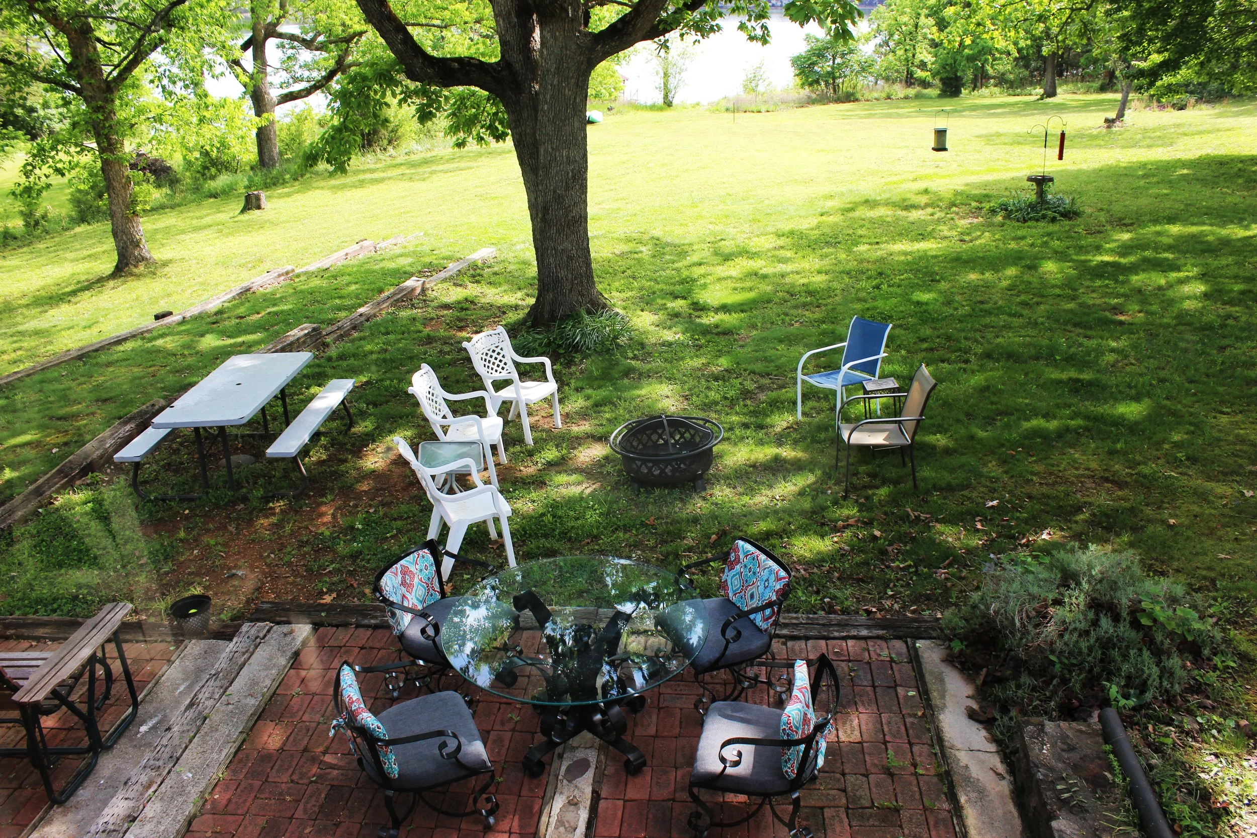 A backyard patio with a round glass table and six chairs, some with floral cushions, on a brick surface under a large tree. There are additional chairs and seating around the patio, and a grassy lawn with trees and garden decorations beyond.