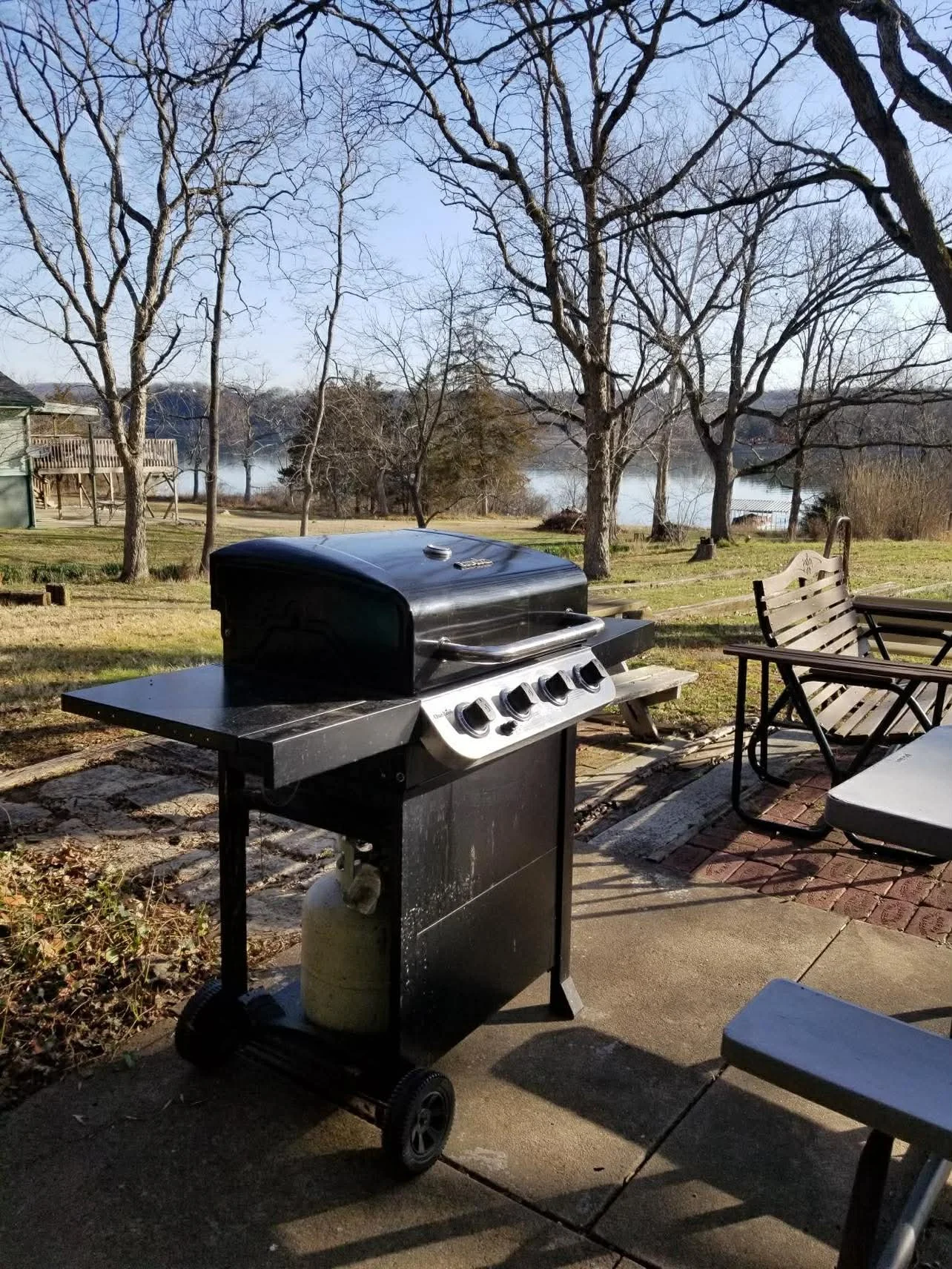 Black gas grill on a patio with outdoor furniture, trees, a lake, and a house in the background.