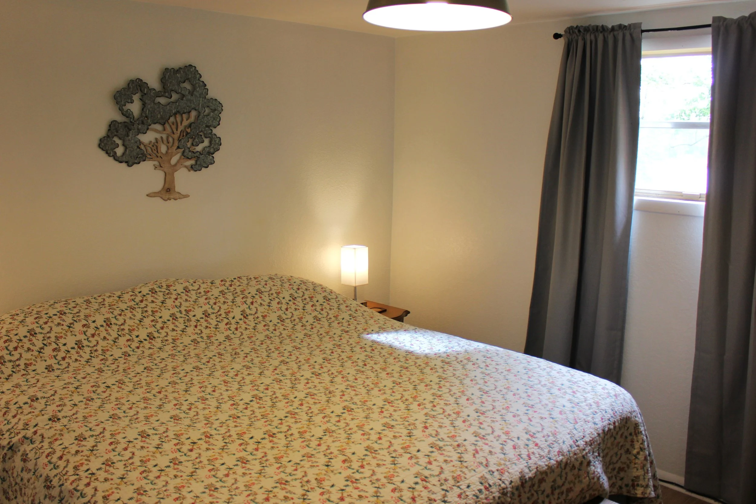A bedroom with a bed covered in a floral patterned quilt, a window with gray curtains, a small bedside table, and a decorative metal tree wall hanging, illuminated by a ceiling light and a small lamp.