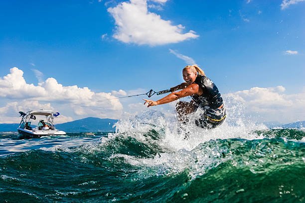 A woman wakeboarding across a large wave on Table Rock Lake, Missouri, with a white motorboat and rolling blue hills in the distance under a bright summer sky.