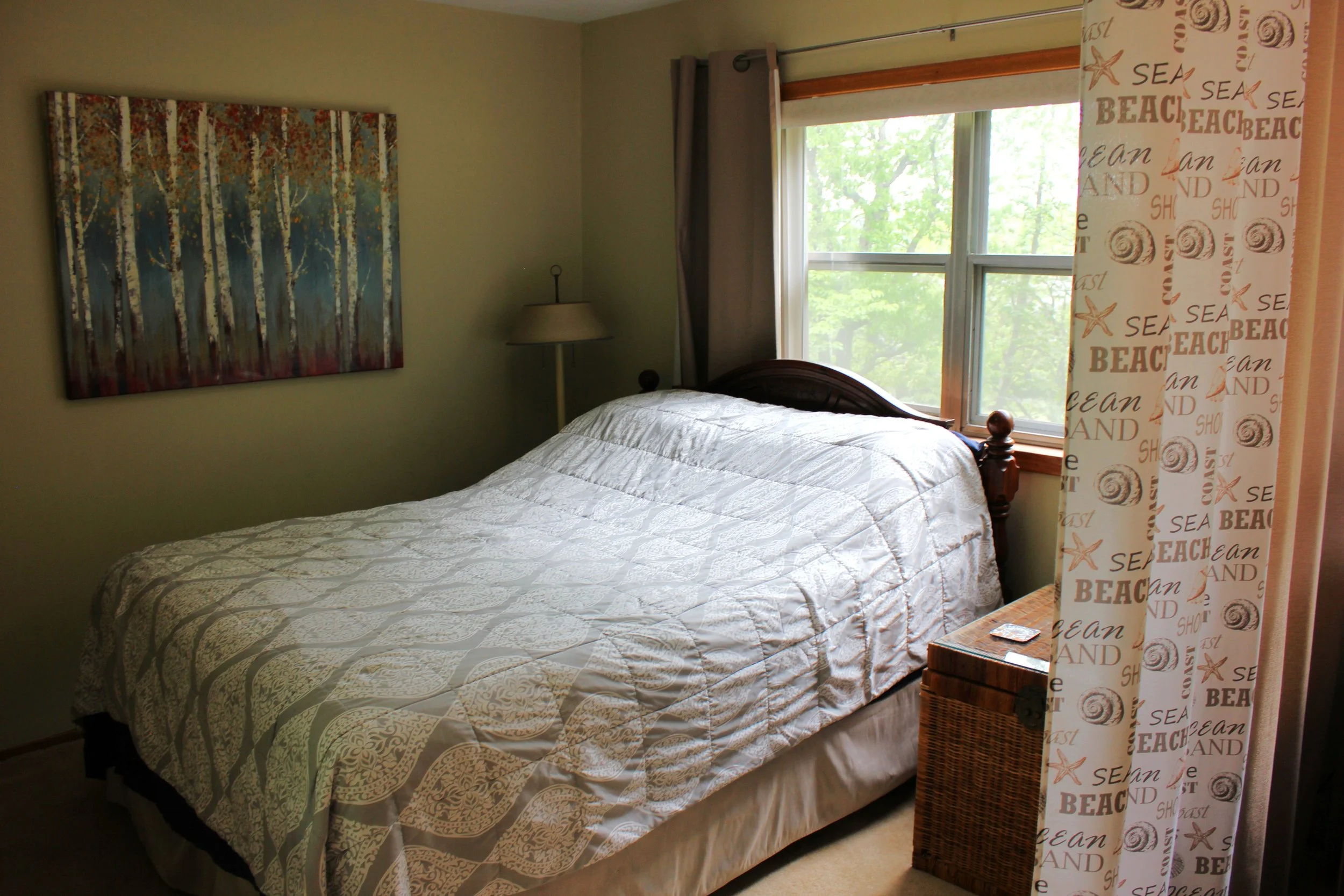 A bedroom with a bed dressed in beige and white patterned bedding, a bedside lamp, artwork of a forest scene, a window with gray curtains, and beach-themed curtains with words like "sea," "beach," and "sand" and starfish and shell designs.