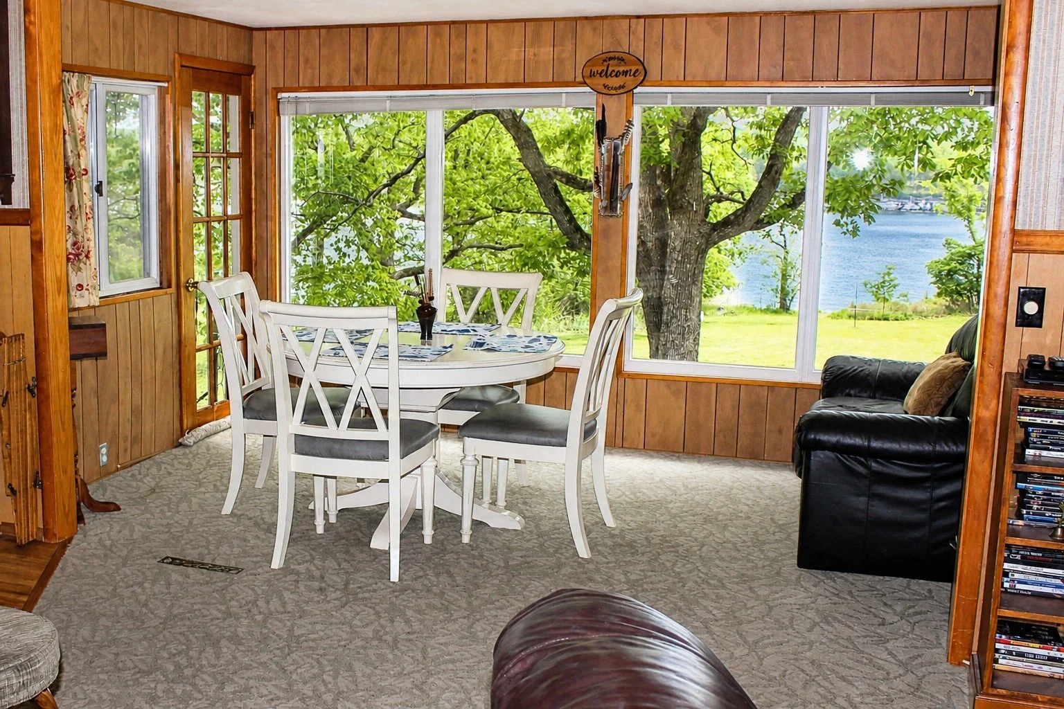 Living room with wood-paneled walls, large windows overlooking a tree and a lake, a white dining table with four chairs, a black leather armchair, and a bookshelf with DVDs.