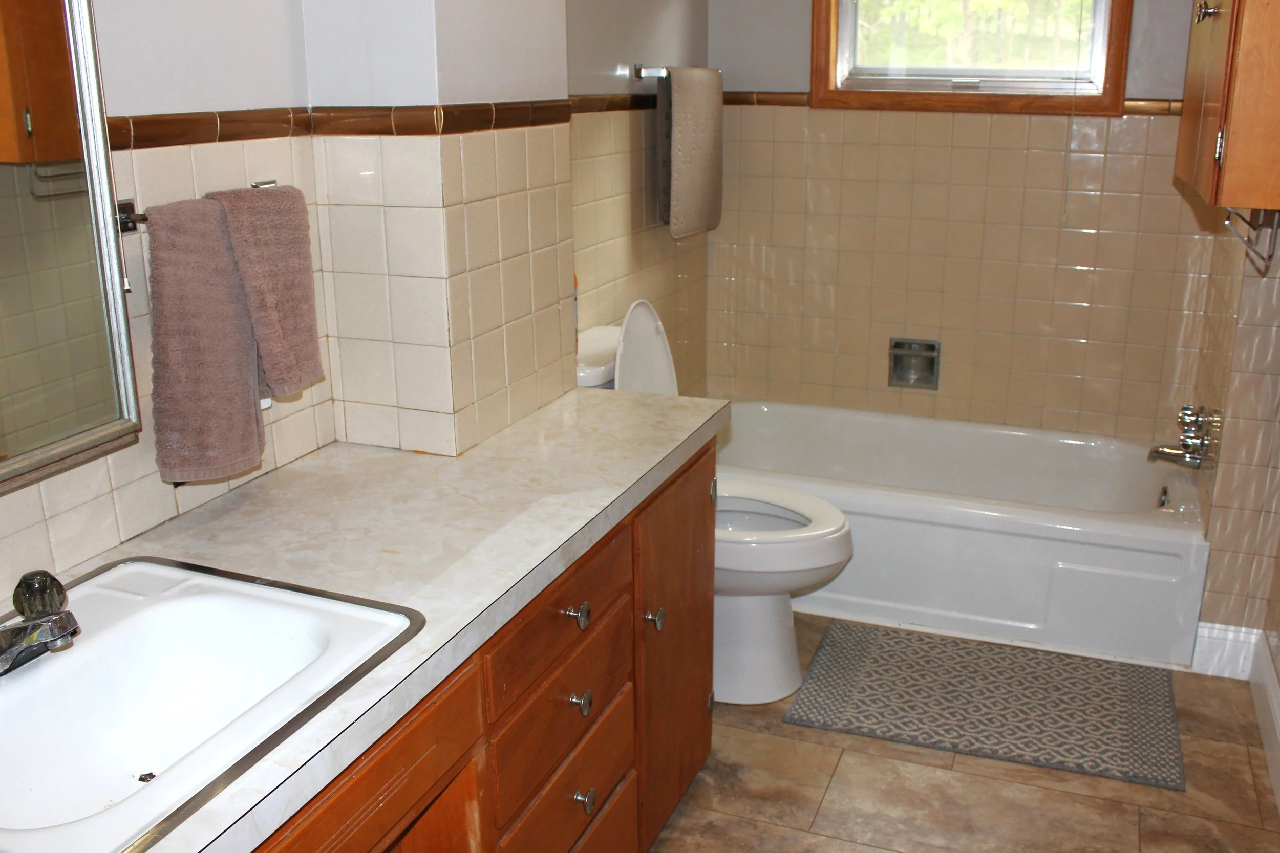 A bathroom with beige tiled walls, a wooden vanity with a cream-colored countertop, a white sink, a toilet, a bathtub, and a window with a wooden frame.