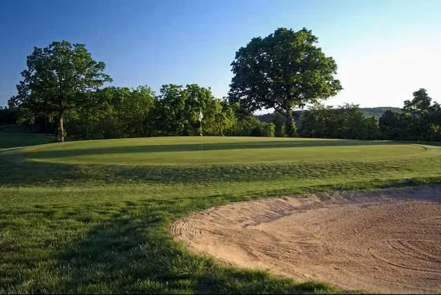 A well-manicured green and sand bunker at the Holiday Island Golf Course near Table Rock Lake, Arkansas, surrounded by lush trees under a clear blue sky.