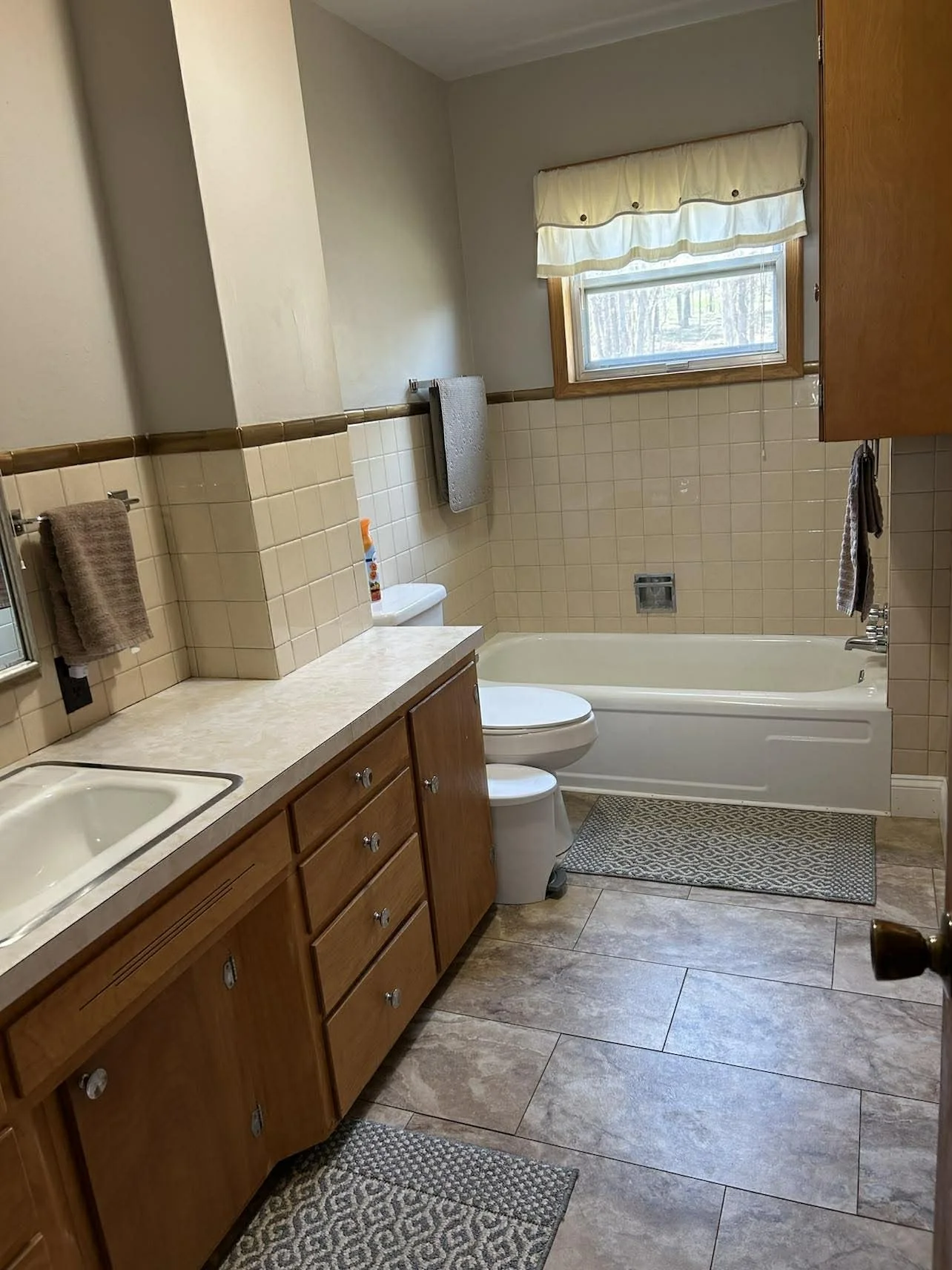 A bathroom with beige tiled walls and floor, a window with a cream valance, wooden cabinets, a white bathtub, toilet, sink, and brown towels.