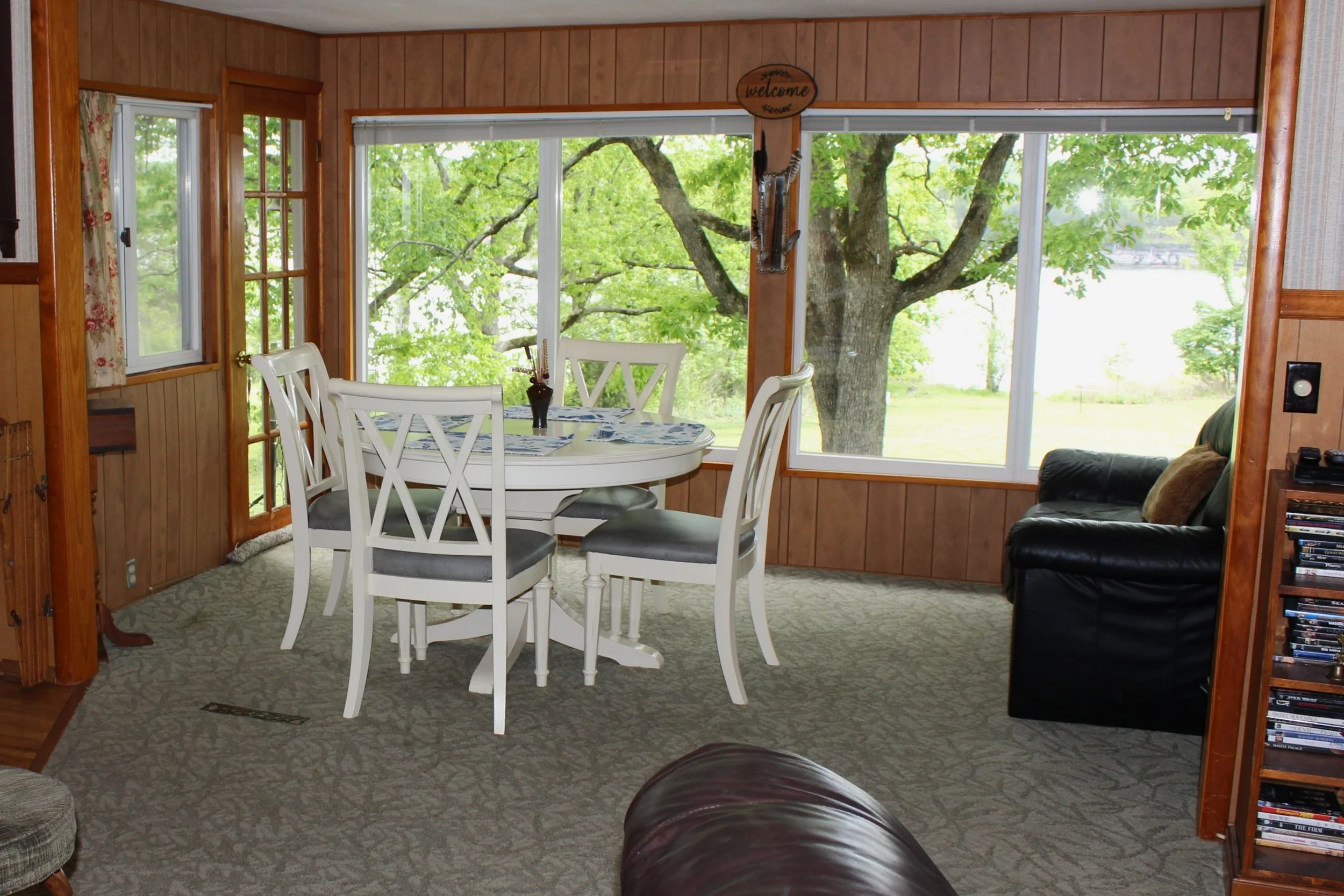 Living room with wood-paneled walls and large windows showing green trees outside. There is a white dining table with four chairs, and a dark leather armchair is partially visible in the foreground.