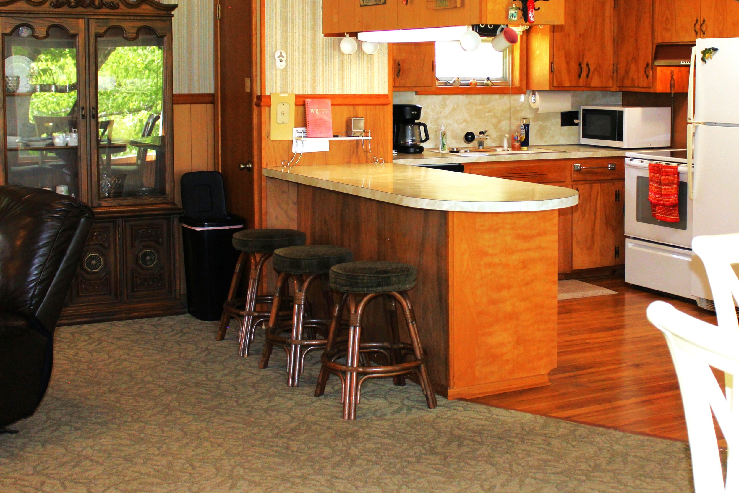 A kitchen with wooden cabinets, a white refrigerator with a red towel, and a countertop with a coffee maker, microwave, and various small items. There are three bar stools with padded seats in front of a curved kitchen island. A glass door with a din
