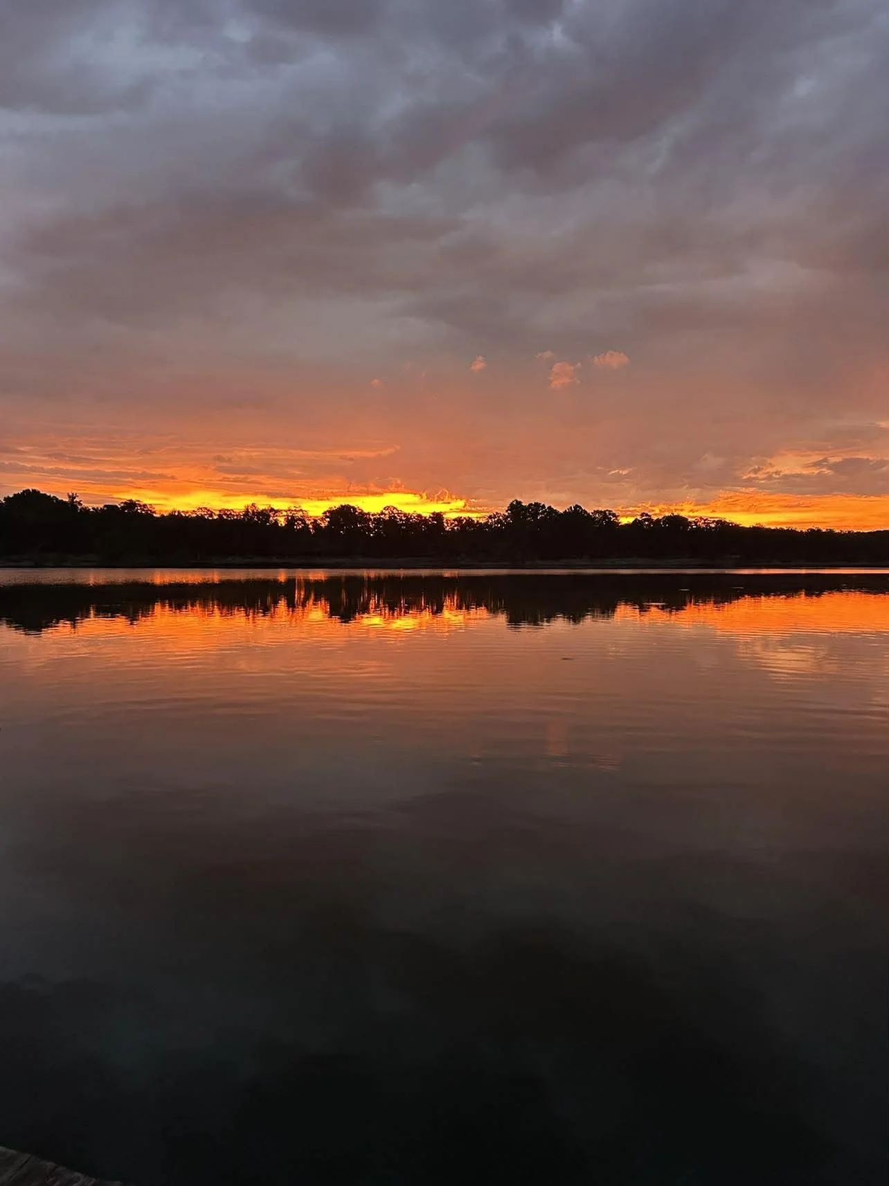 Sunset over a calm body of water with a partly cloudy sky and silhouetted trees in the distance.