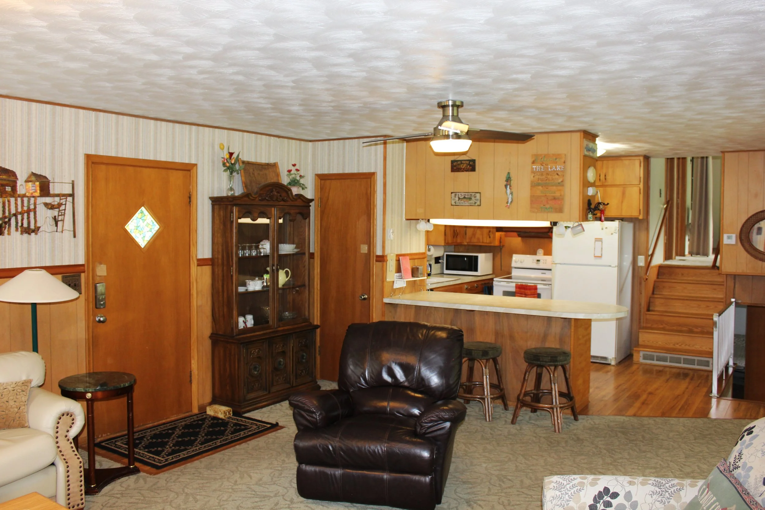 Living room with leather armchair, beige and floral armchair, small side tables, wooden cabinet with glass doors, wall decorations, and a view into the kitchen area with white appliances and wooden cabinetry.