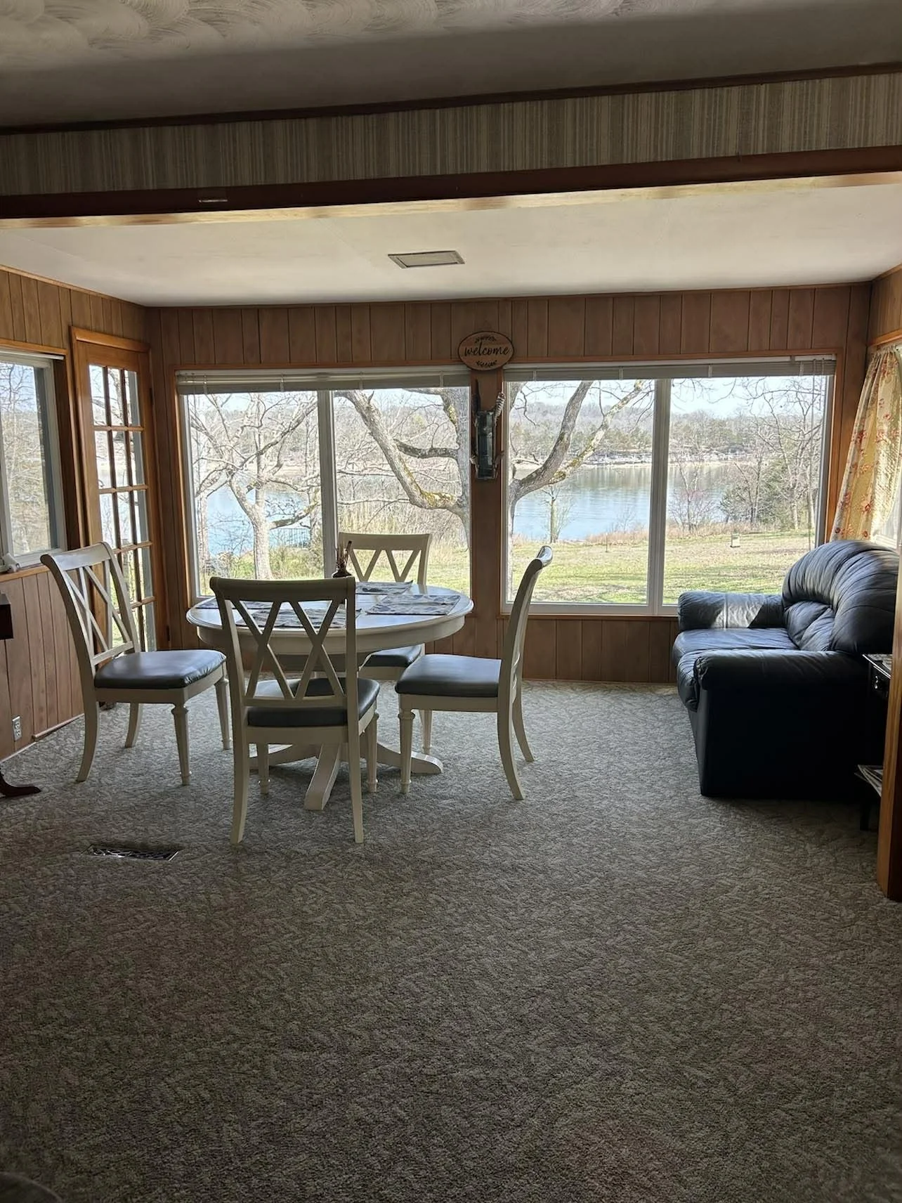 Living room with large windows overlooking Table Rock Lake, with a round dining table and four chairs, a black leather sofa, and wooden wall paneling.