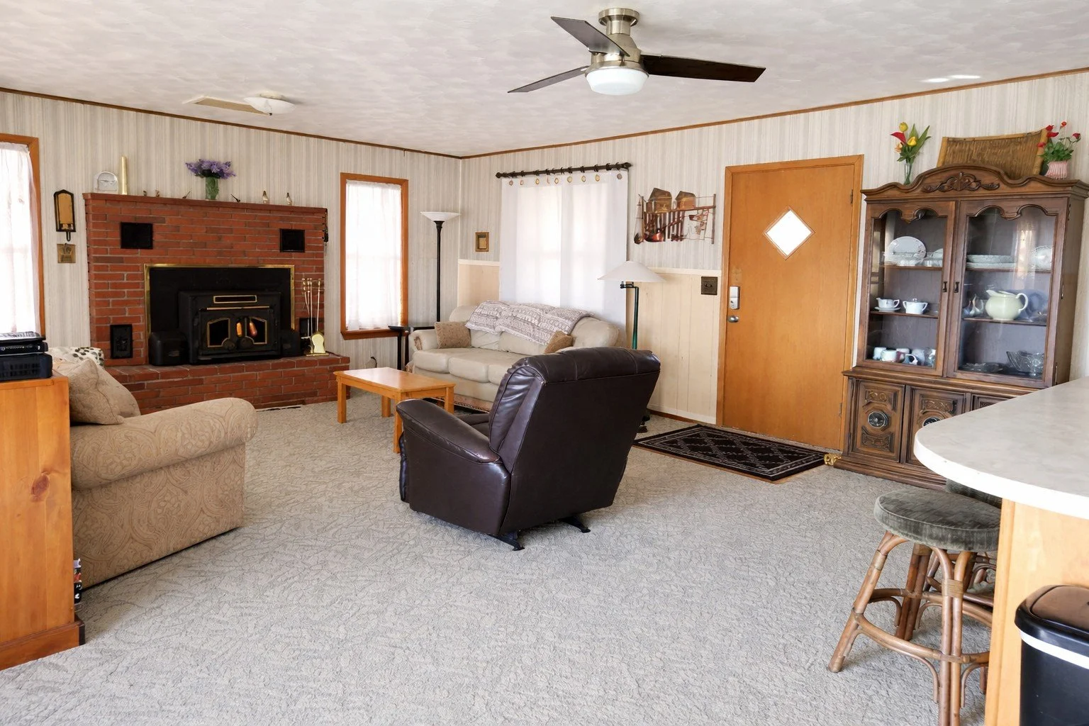 Living room with beige carpet, brick fireplace, beige and black sofas, wooden coffee table, upright and floor lamps, wood-paneled walls, white curtains, wooden door, glass display cabinet with china, and a ceiling fan.