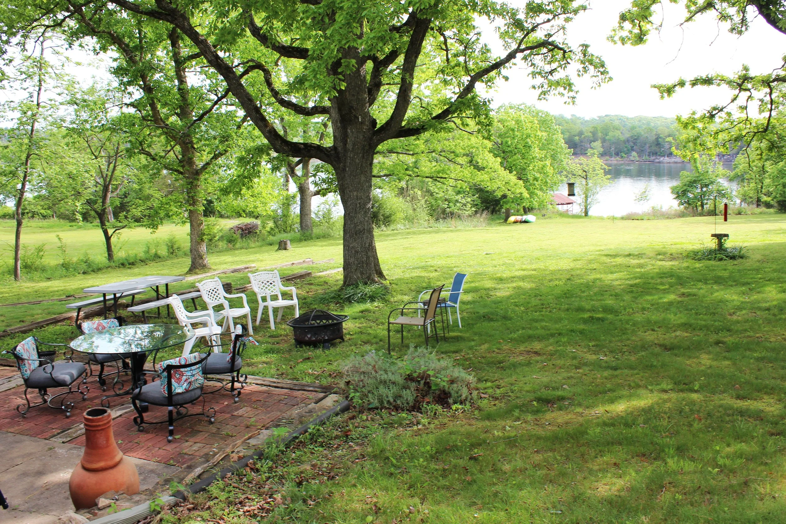 A backyard with a large tree and a view of a lake in the background. There are several chairs and tables on a brick patio and a grassy lawn.