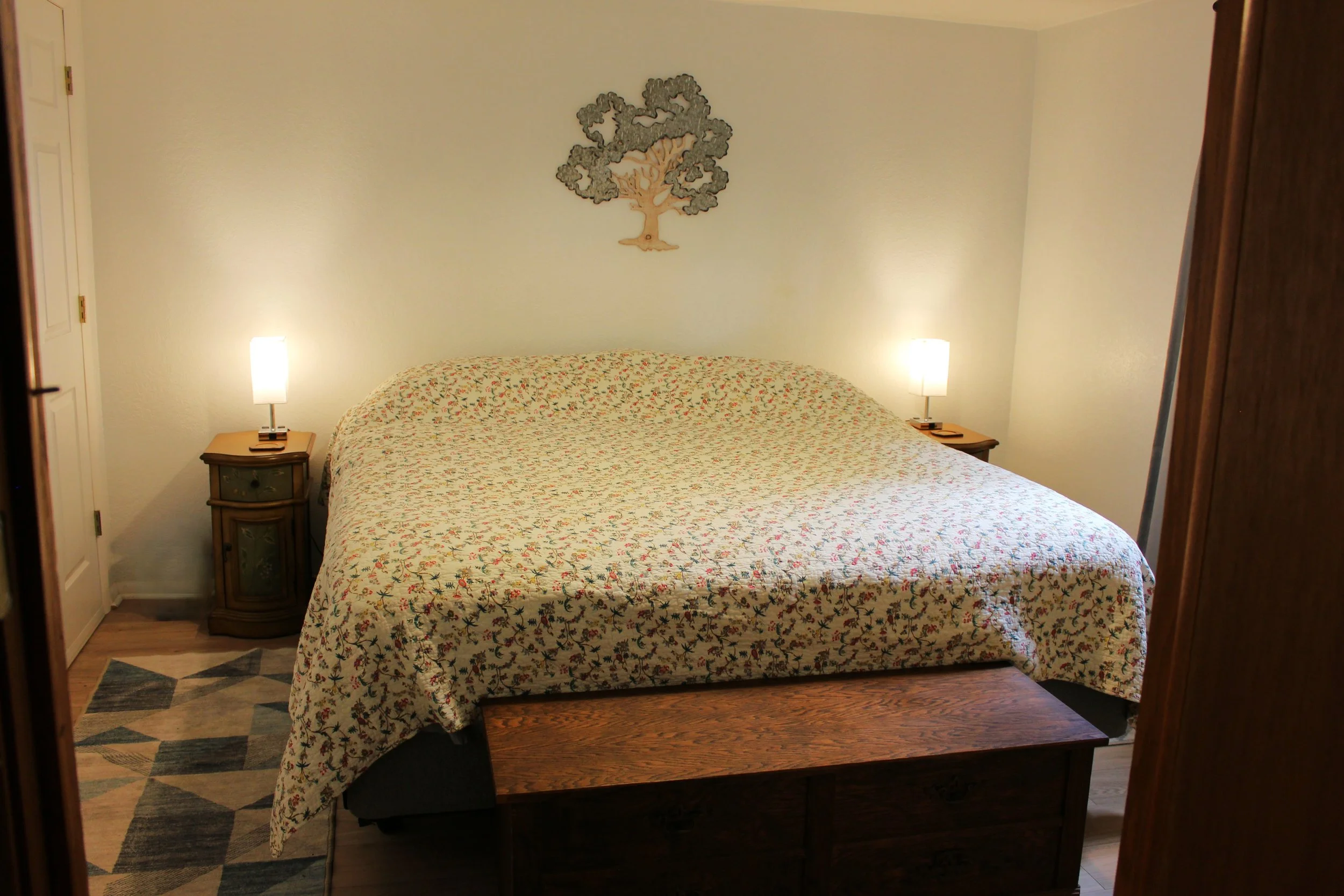 A bedroom with a bed covered in a floral quilt, two bedside tables with lamps, a wall decoration of a tree, and part of a geometric rug on a wooden floor.