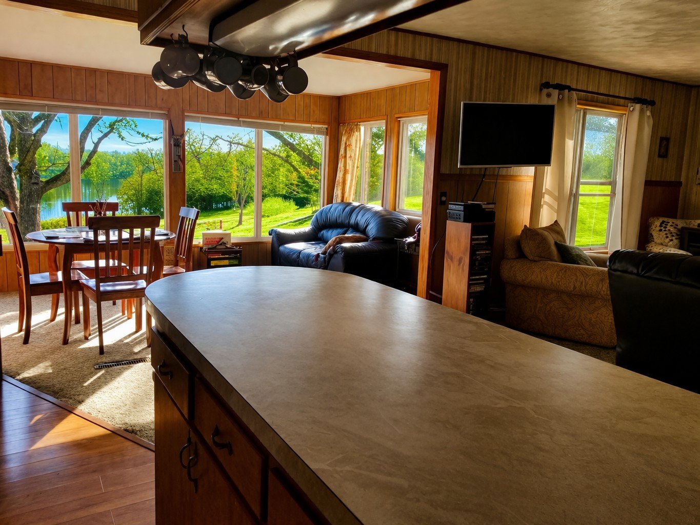 Living room with large windows overlooking a green outdoor landscape, a dining table with chairs, a black leather sofa, a patterned armchair, and a mounted flat-screen TV.