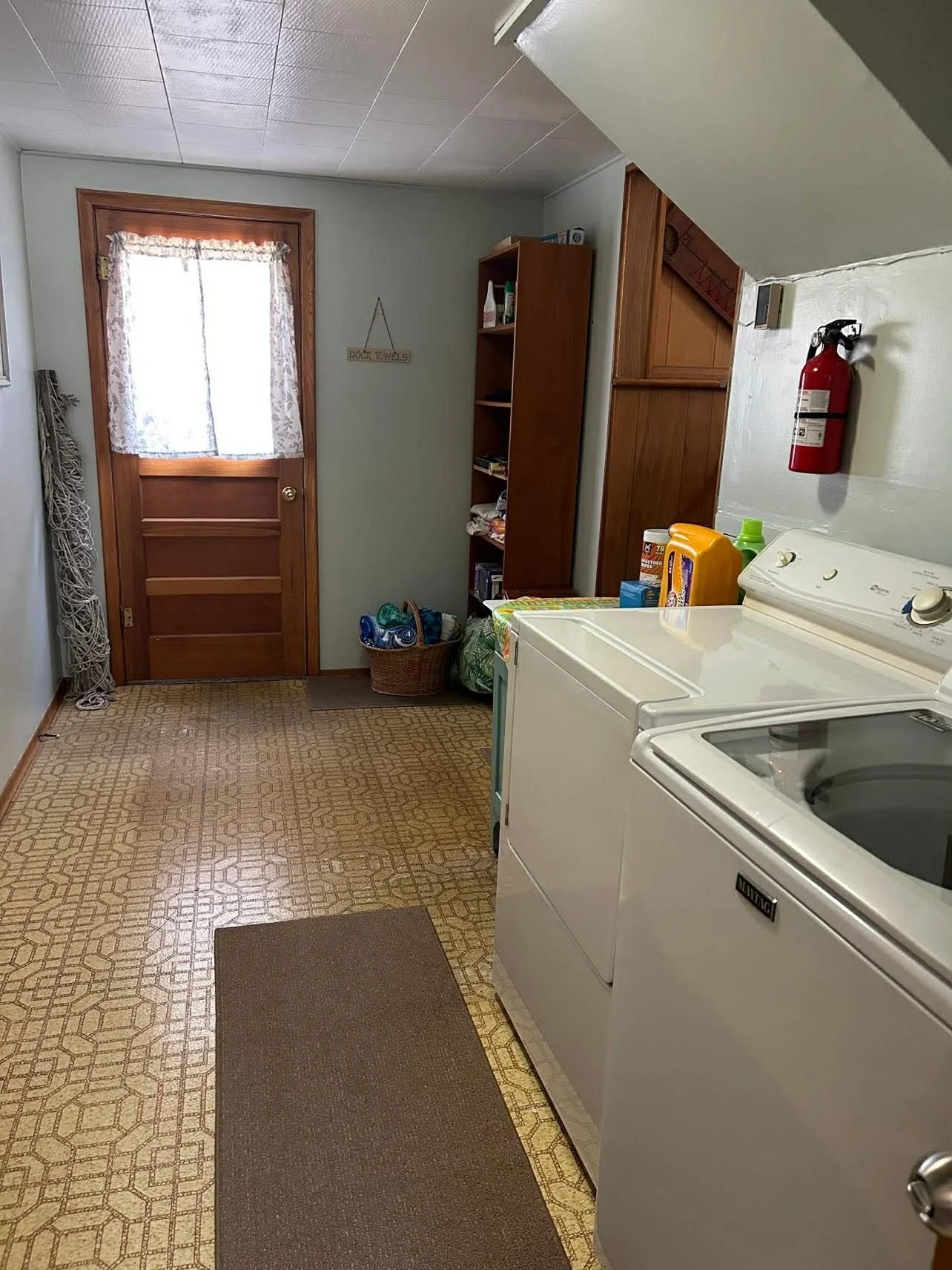 Laundry room with washing machine, shelf with cleaning supplies, fire extinguisher on wall, door with lace curtains, laundry basket, and a brown rug on patterned yellow floor.