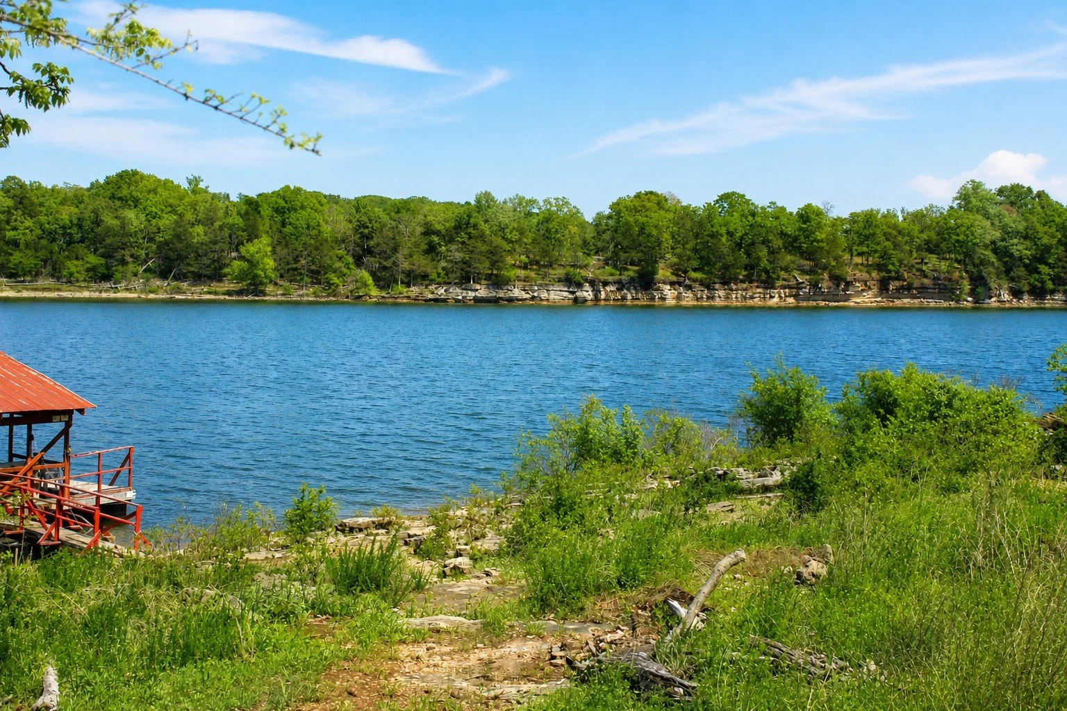 View of a lake with forested shoreline, blue sky with clouds, and a small red wooden structure near the water's edge surrounded by green grass and shrubs.