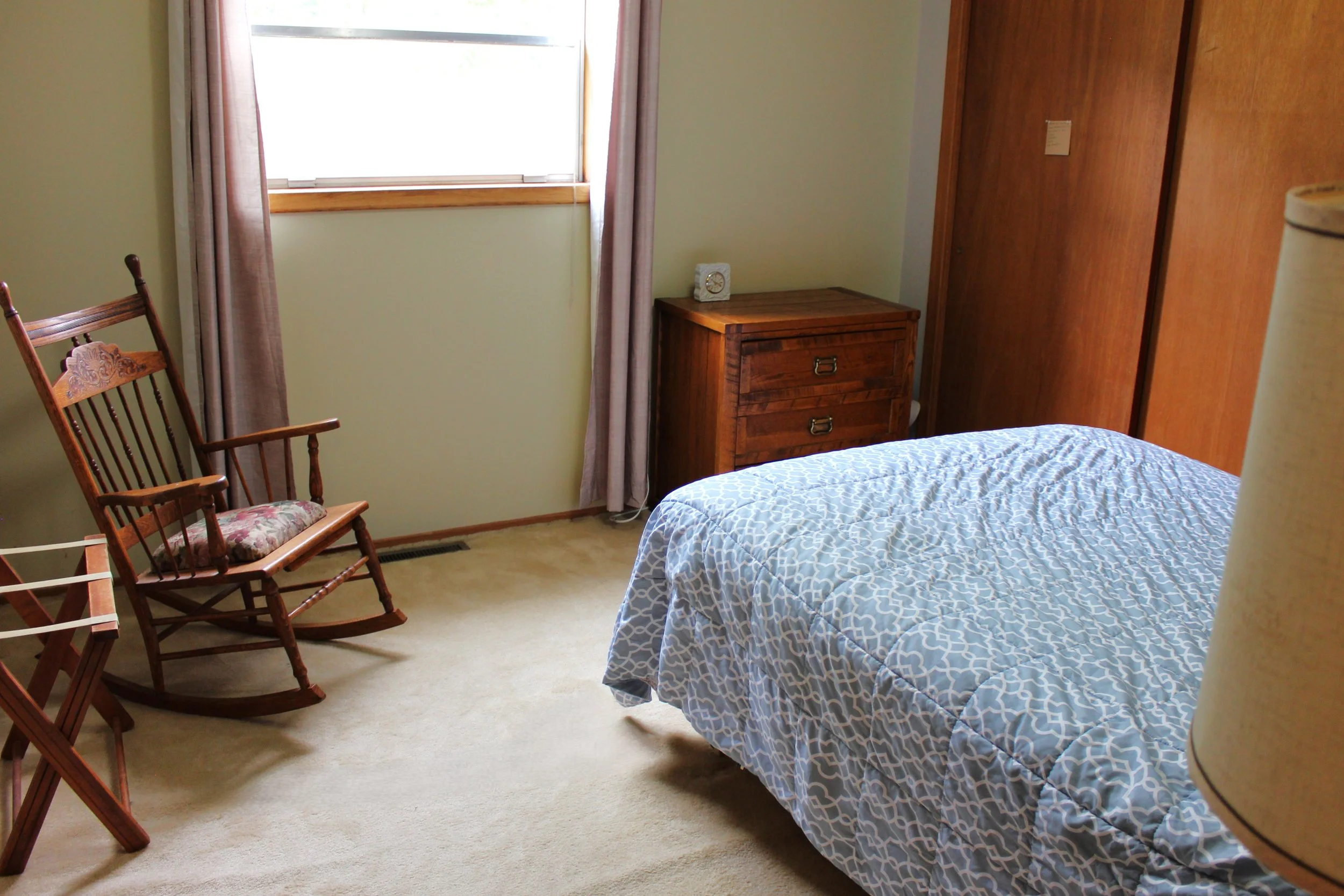 A bedroom with a bed covered in a blue and white geometric patterned comforter, a wooden nightstand with a clock, wooden sliding closet doors, a wooden rocking chair with a floral cushion, a window with pink curtains, and beige carpeted flooring.