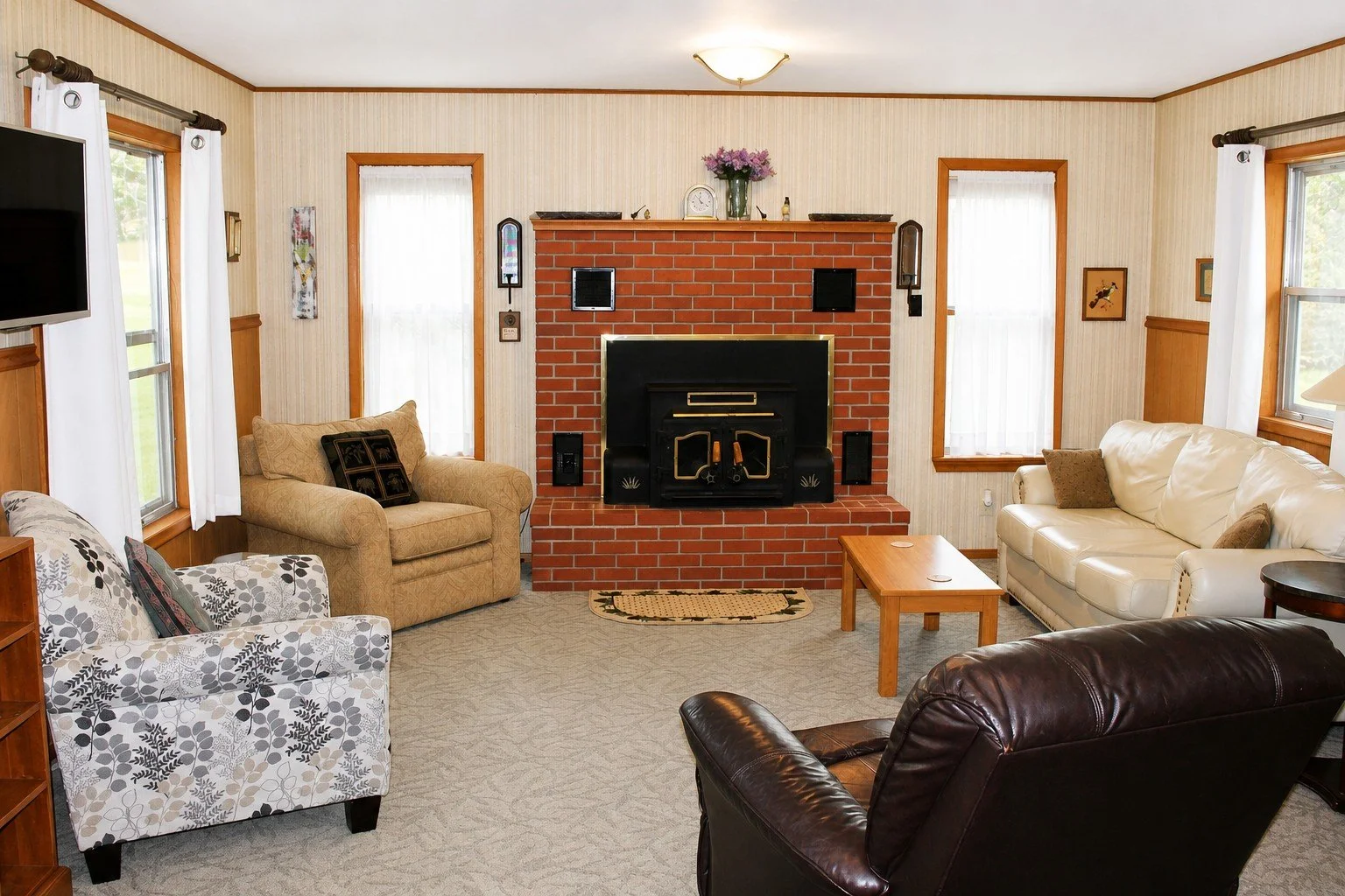 Living room with beige, white, and brown sofas surrounding a brick fireplace, with windows on either side, and a small wooden coffee table in front.