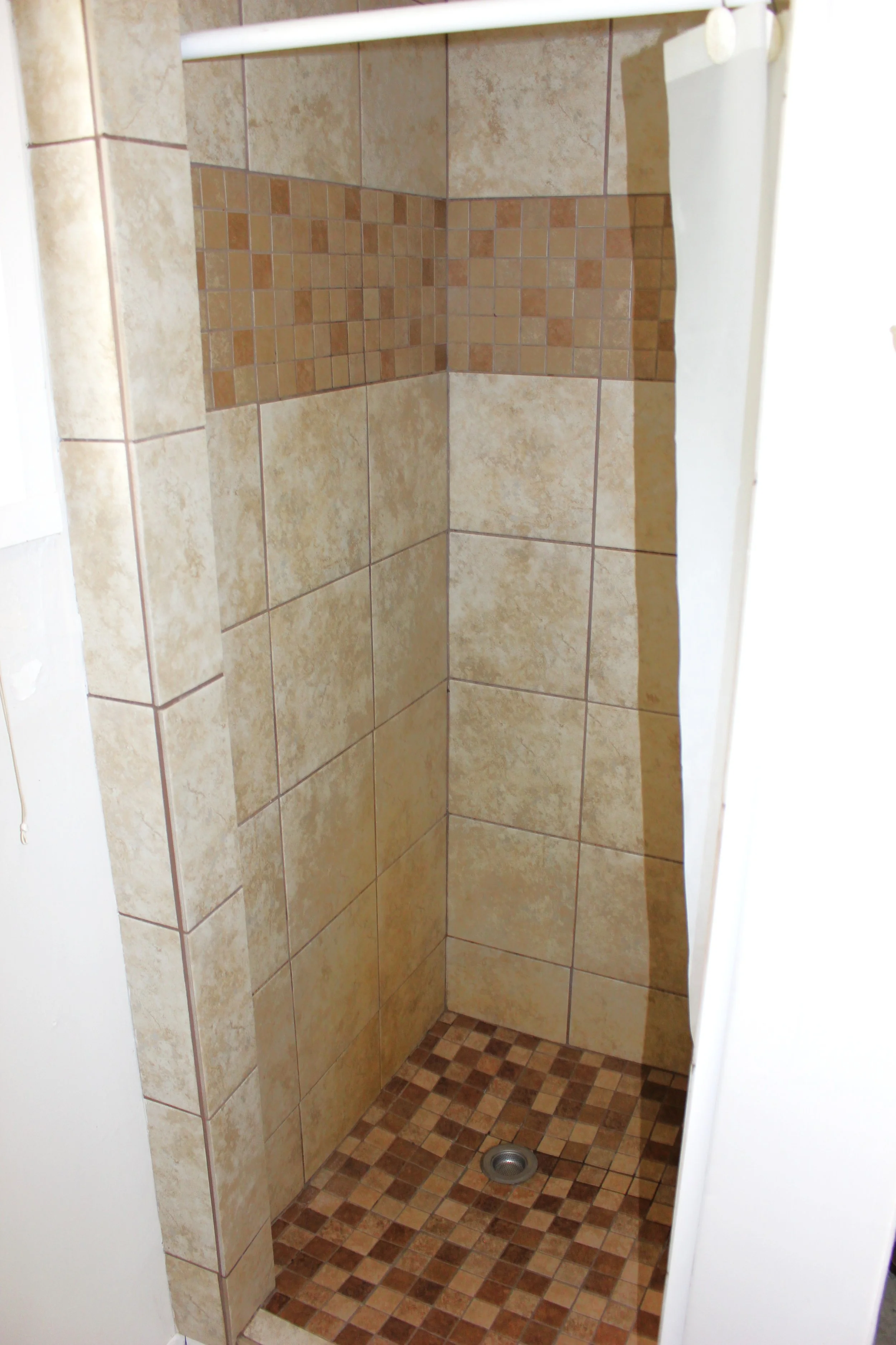 Corner of a shower with beige and brown tiled walls and a small brown tiled floor.