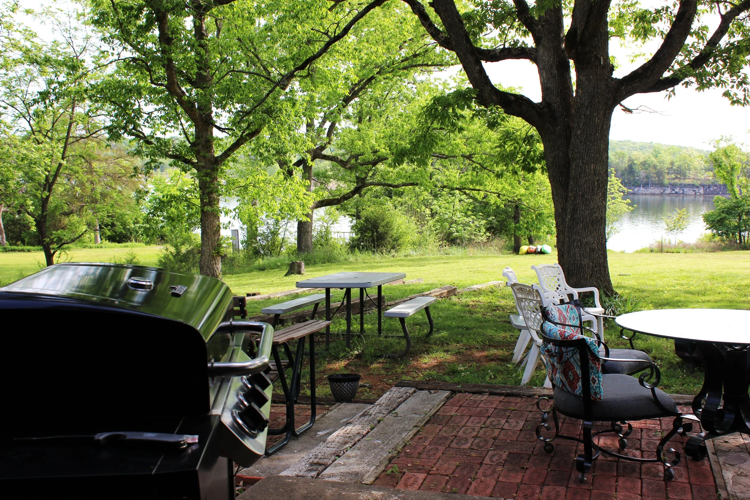 A backyard patio scene with a barbecue grill in the foreground, outdoor tables and chairs, green trees, and a body of water in the background, indicating a peaceful outdoor setting on a sunny day.