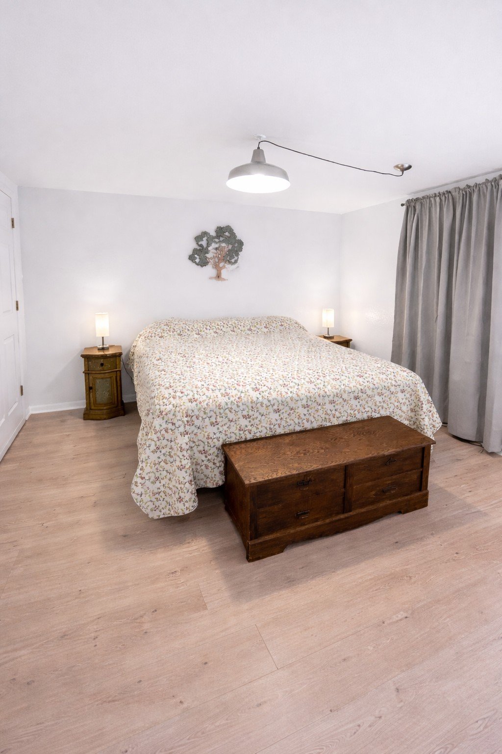 A bedroom with a bed covered in a light patterned quilt, two bedside tables with lamps, a wooden chest at the foot of the bed, gray curtains, and a decorative wall piece above the bed.